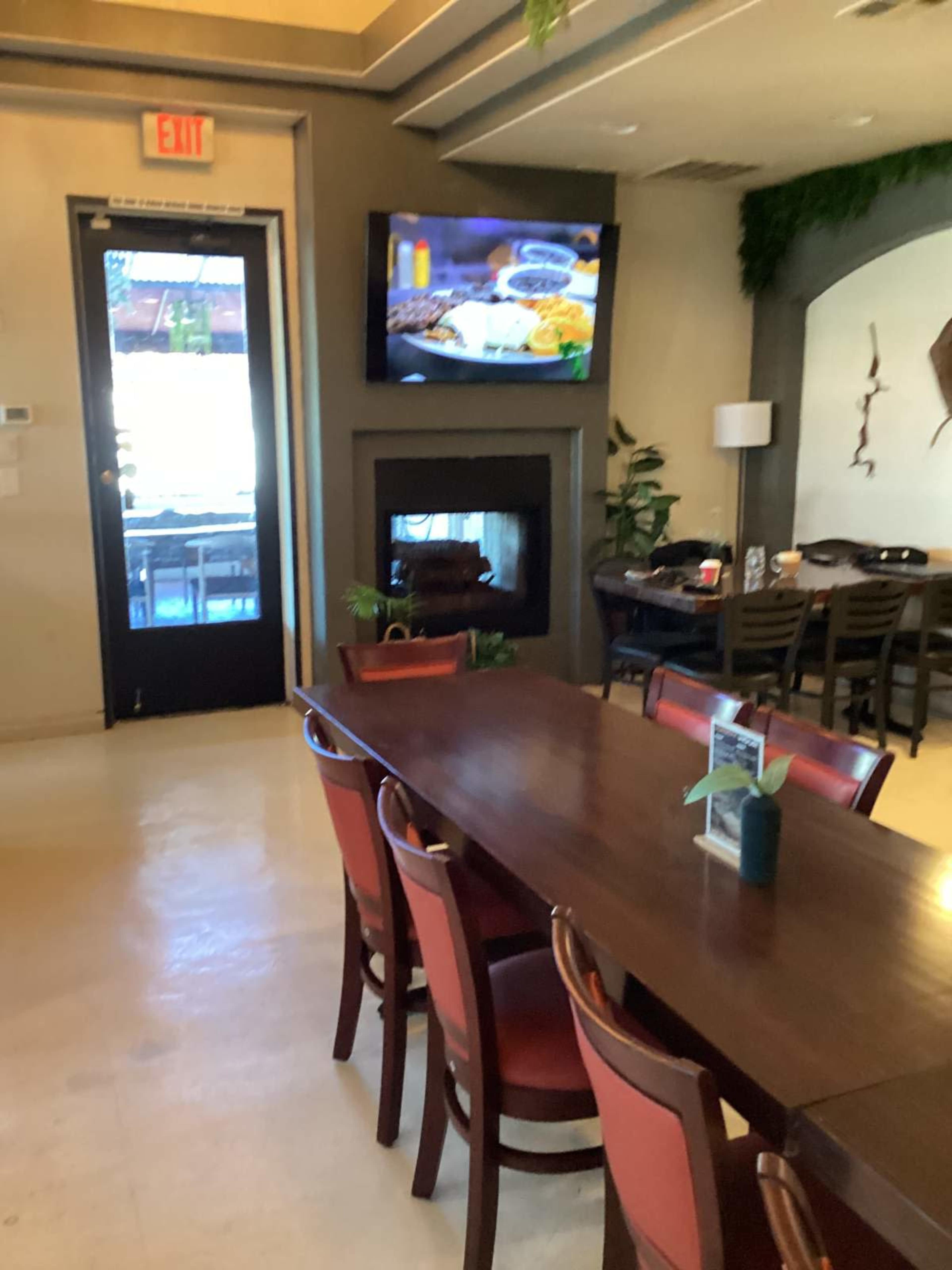 The image shows an interior view of a restaurant with a long wooden table surrounded by chairs, a television displaying food, and a fireplace.