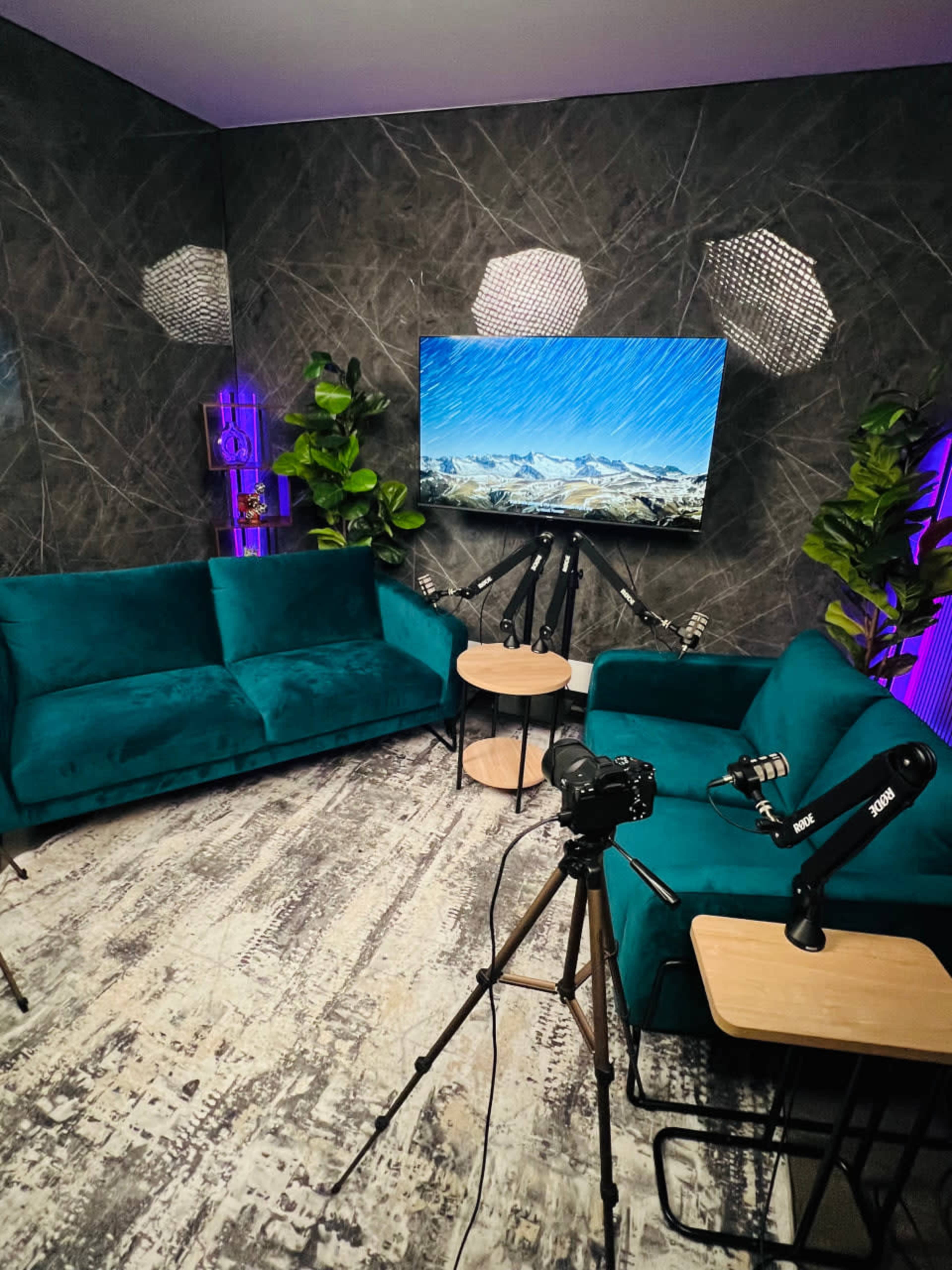 The image shows a modern seating area with two teal sofas, a round table, and a camera setup facing a large screen displaying a mountain landscape.