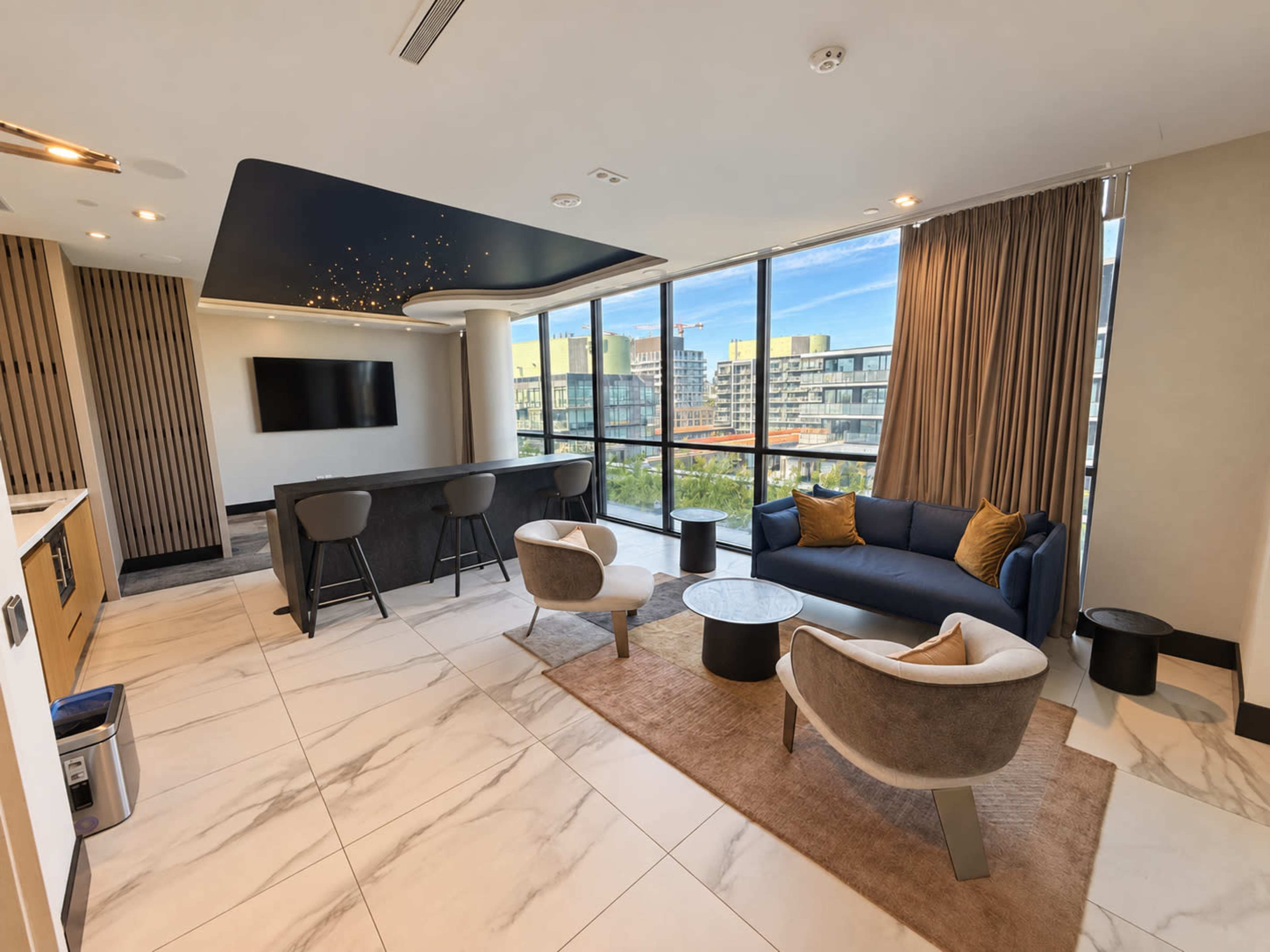 A modern living space features a blue couch, two sculptural chairs, and a counter with bar stools, all illuminated by natural light from large windows.