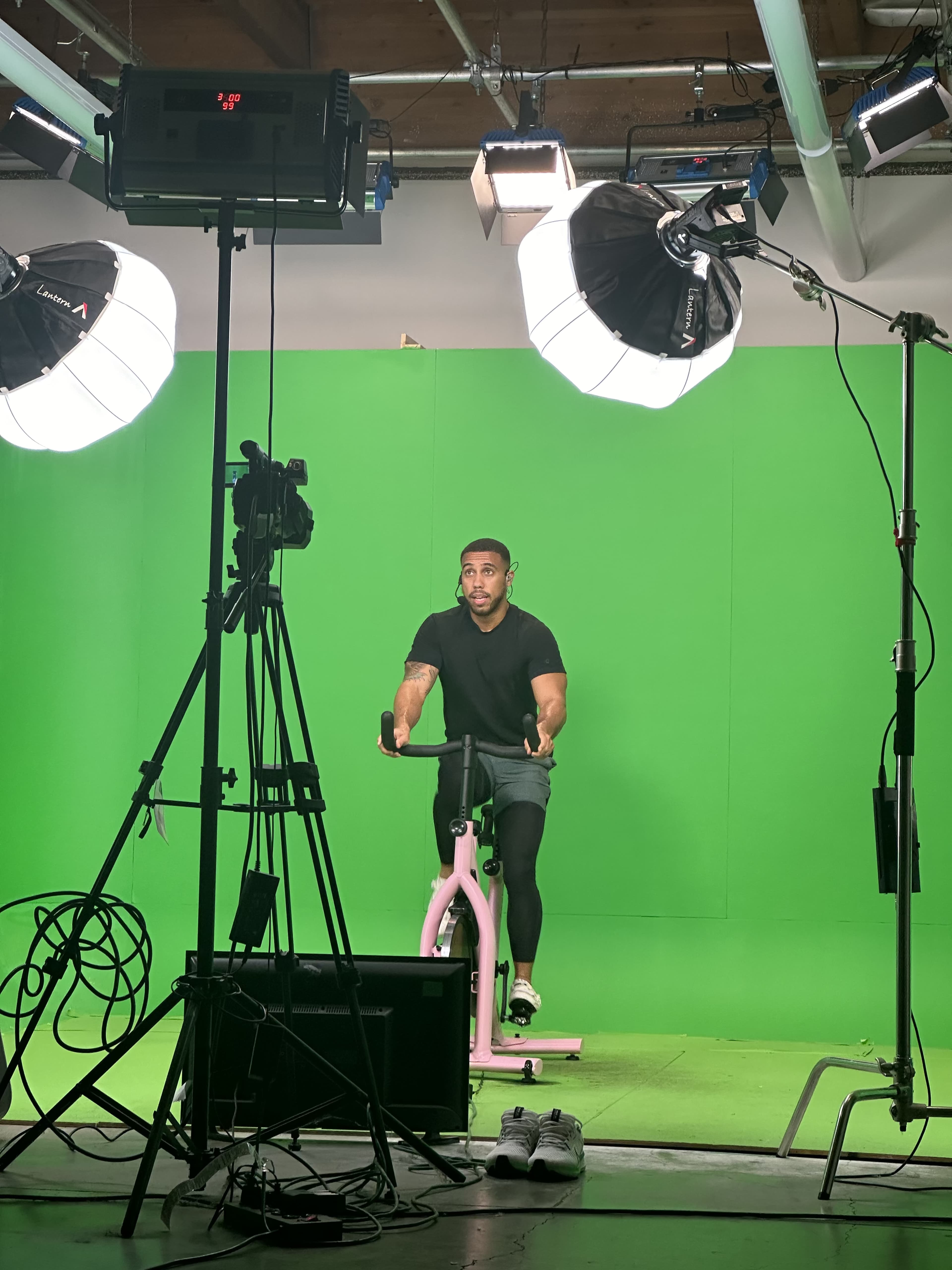 A man is cycling on a stationary bike in front of a green screen, with studio lights and cameras positioned around him.