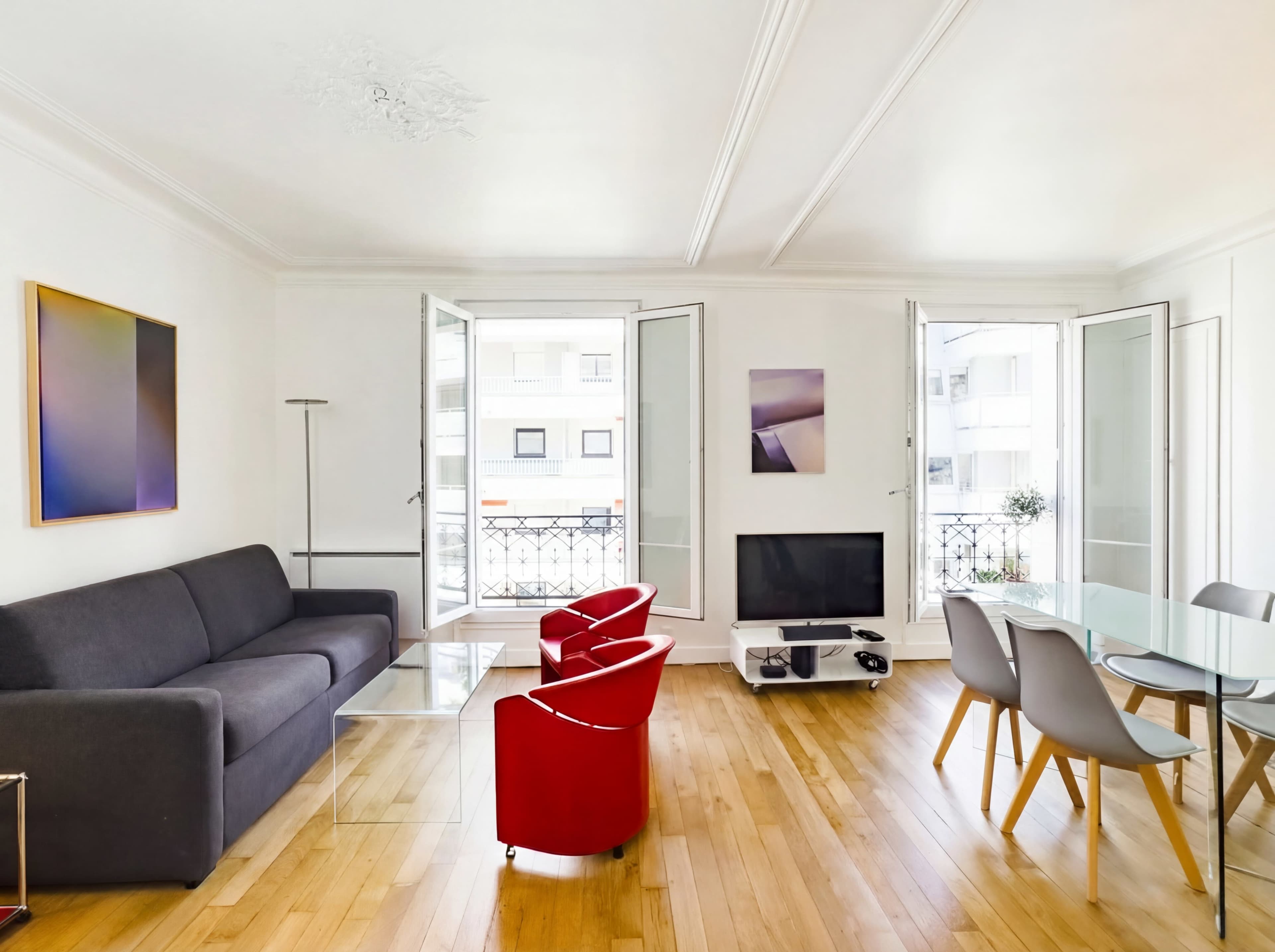 The image features a modern living room with large windows, a gray sofa, red chairs, a glass dining table, and a television against a white wall.