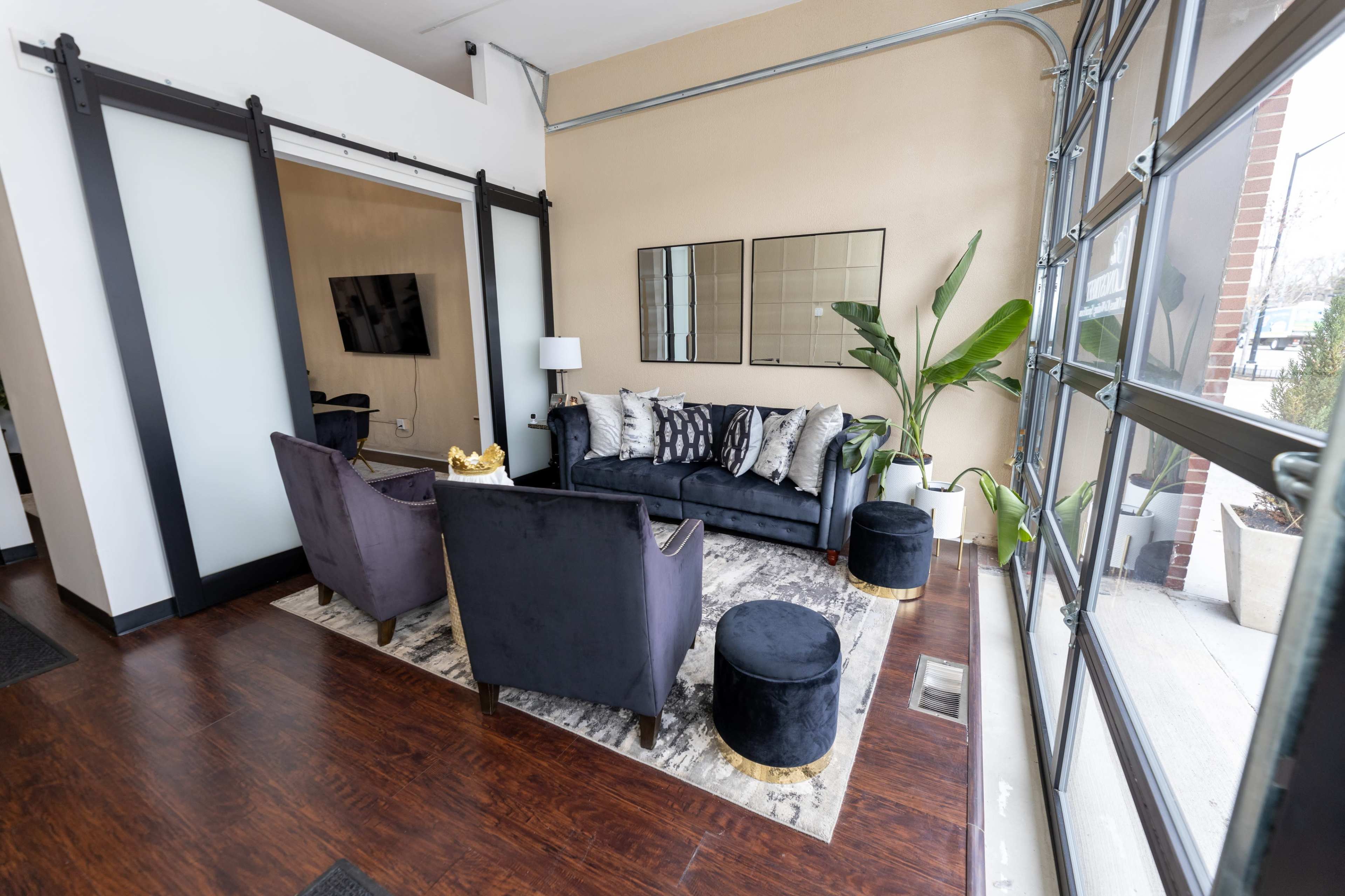 A modern living space features a dark sofa, two armchairs, and decorative pillows on a patterned rug, with a large window providing natural light.