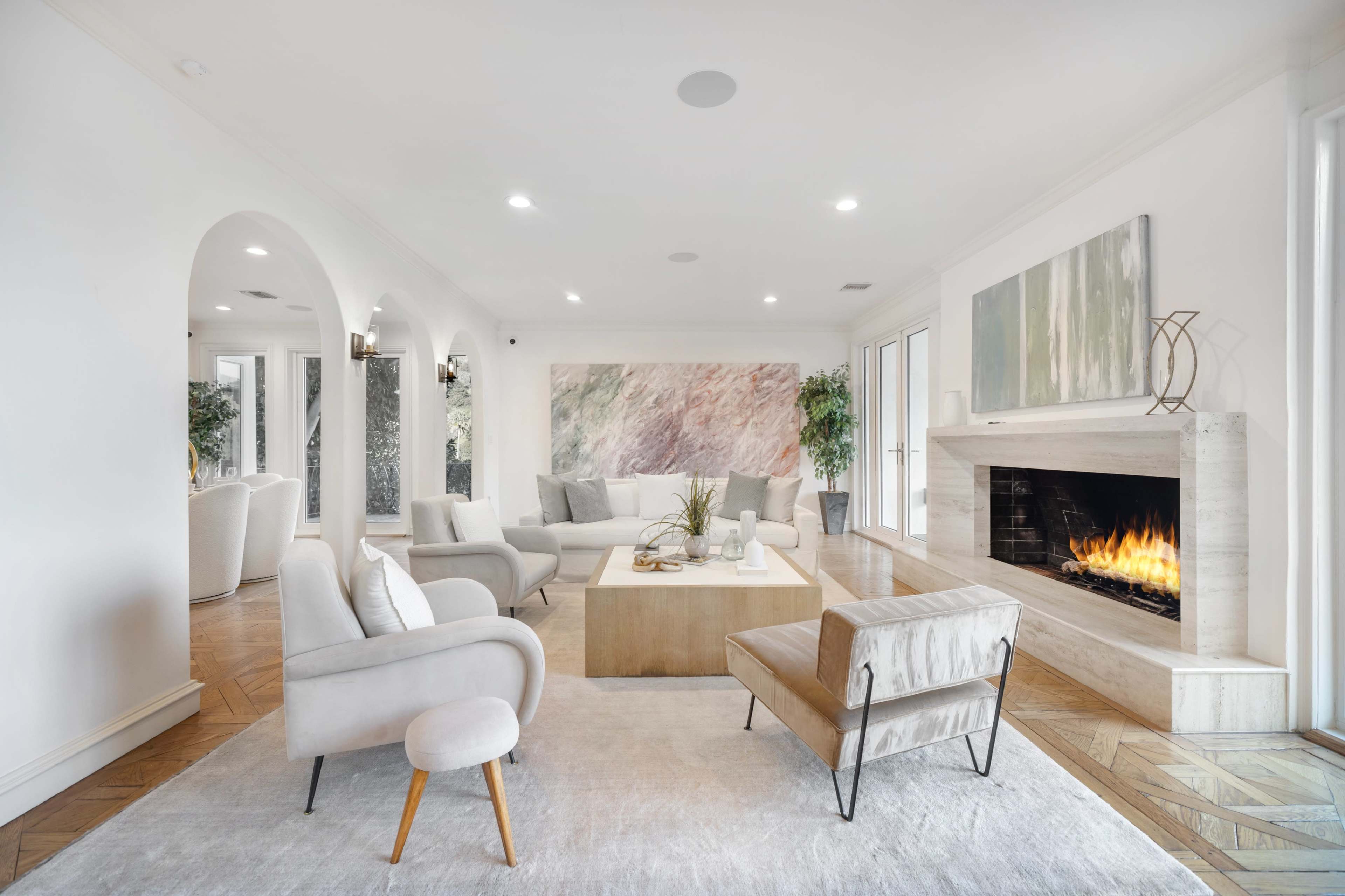 The image shows a spacious modern living room with a stone fireplace, light-colored furniture, and large windows that provide abundant natural light.