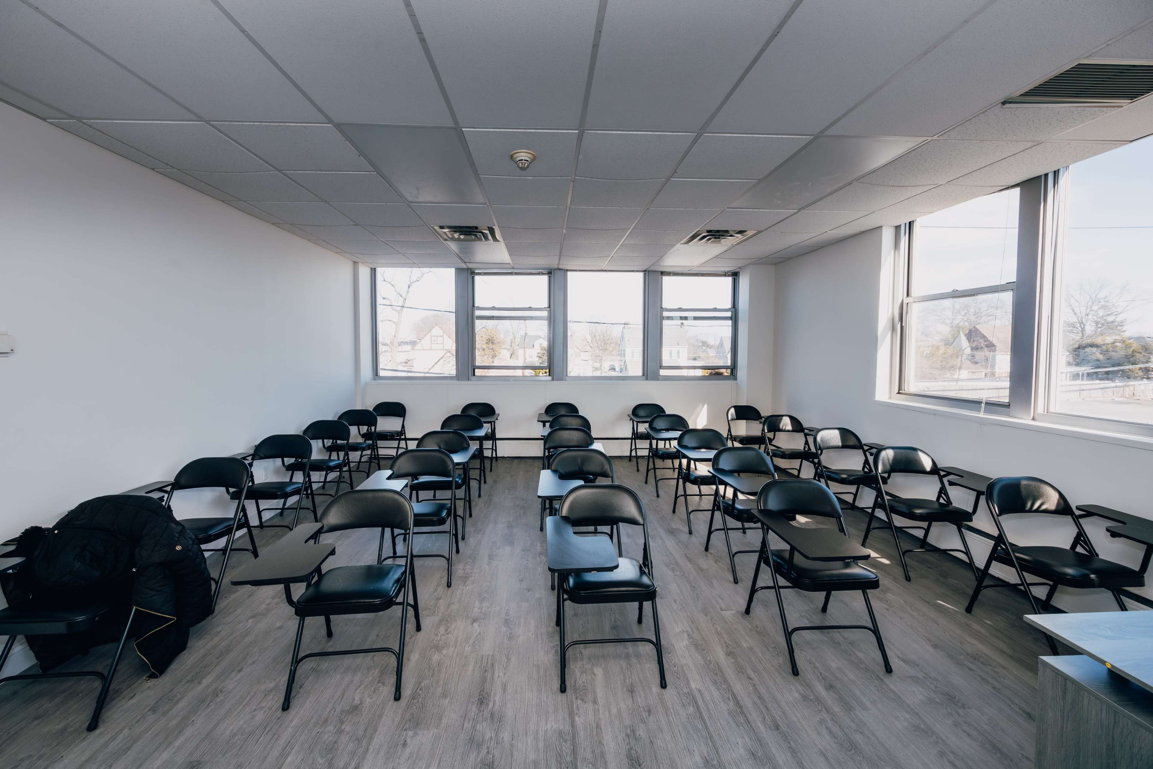 The image shows a nearly empty room with rows of black chairs arranged in neat rows and large windows letting in natural light.
