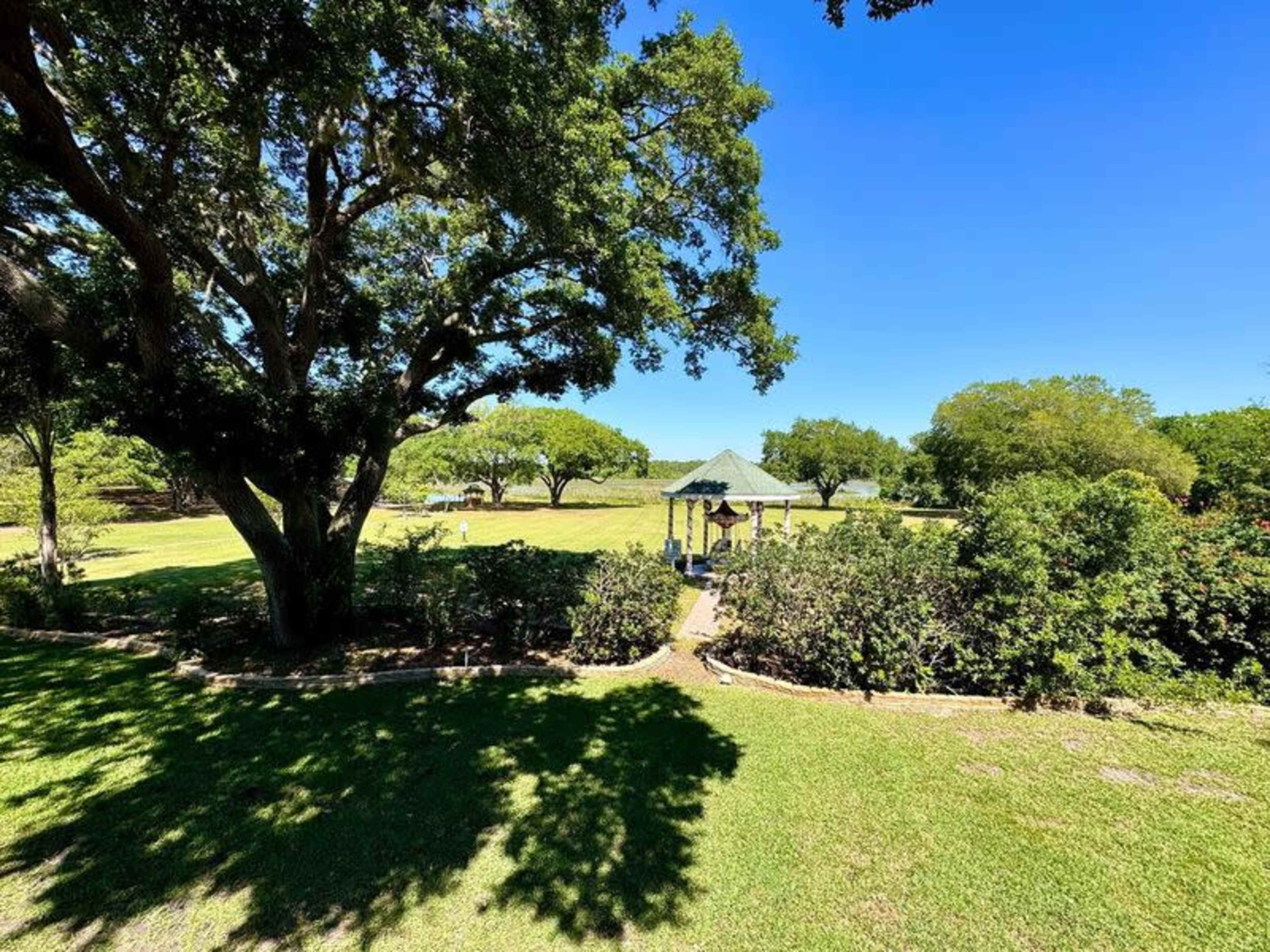 A gazebo is situated in a grassy park surrounded by trees and bushes under a clear blue sky.