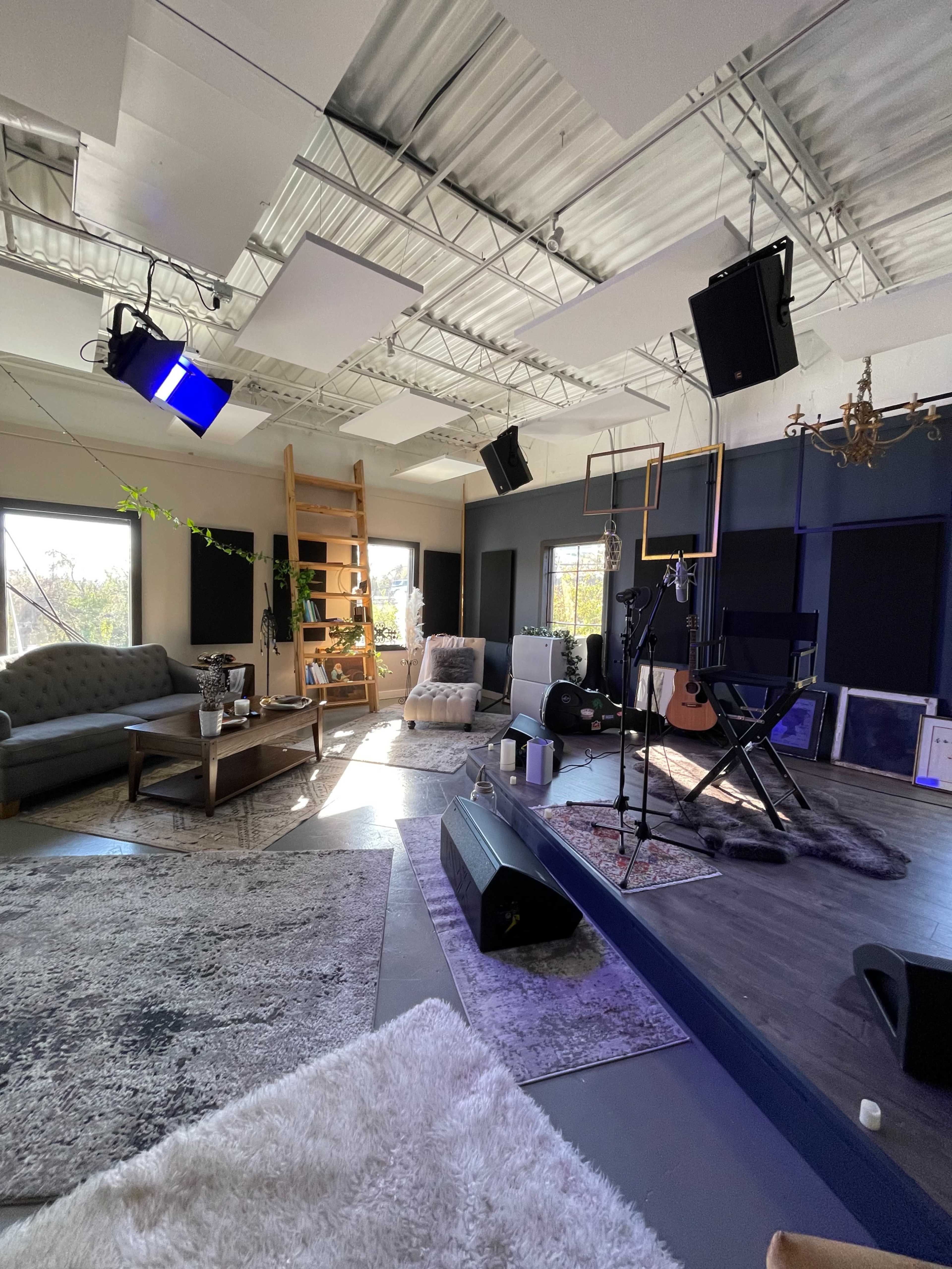 The image shows a well-decorated recording studio featuring a lounge area with a couch, a wooden ladder, audio equipment, and musical instruments arranged on the floor.