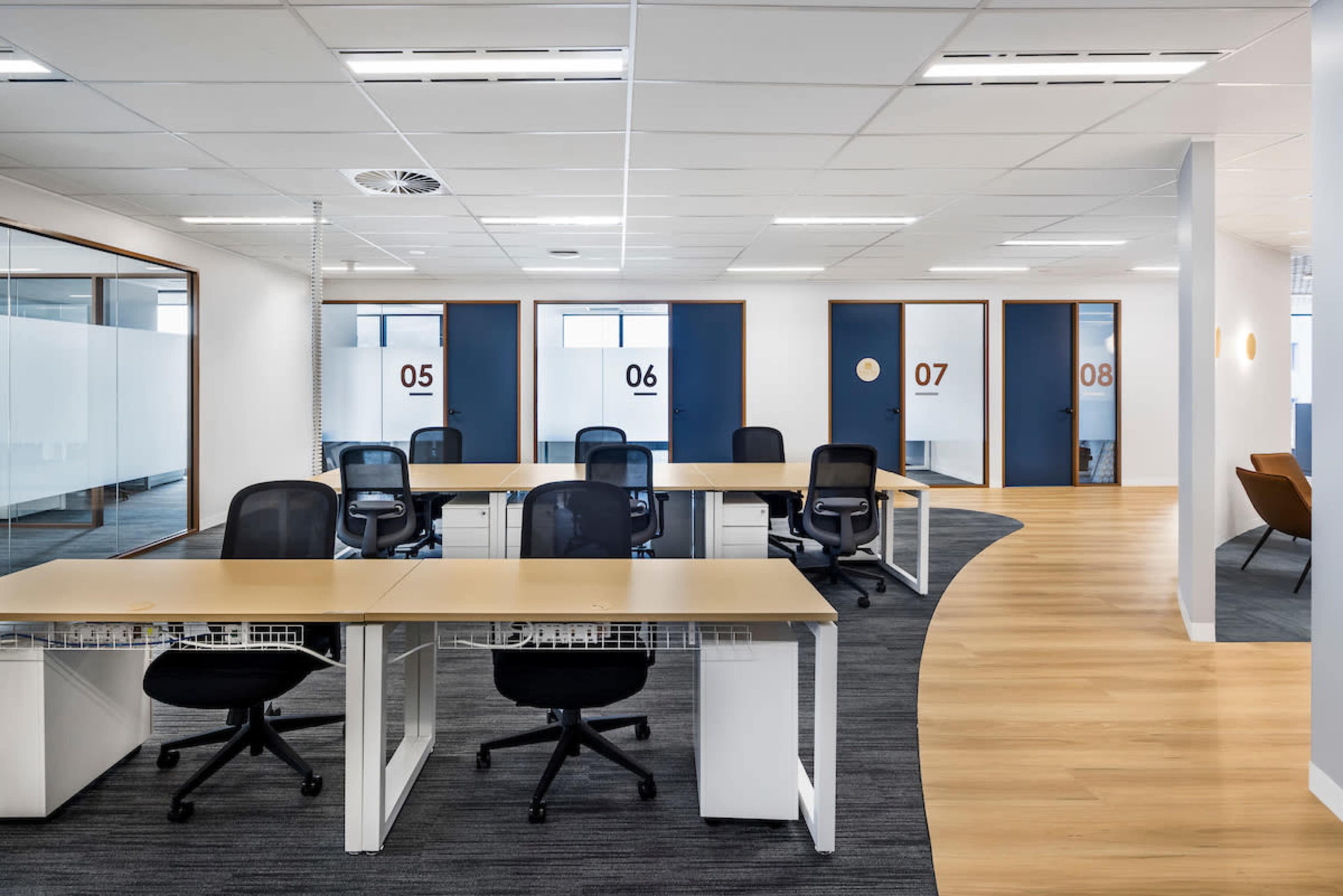 The image shows a modern office space with several empty desks arranged in an open area, surrounded by closed meeting room doors numbered 05, 06, 07, and 08.