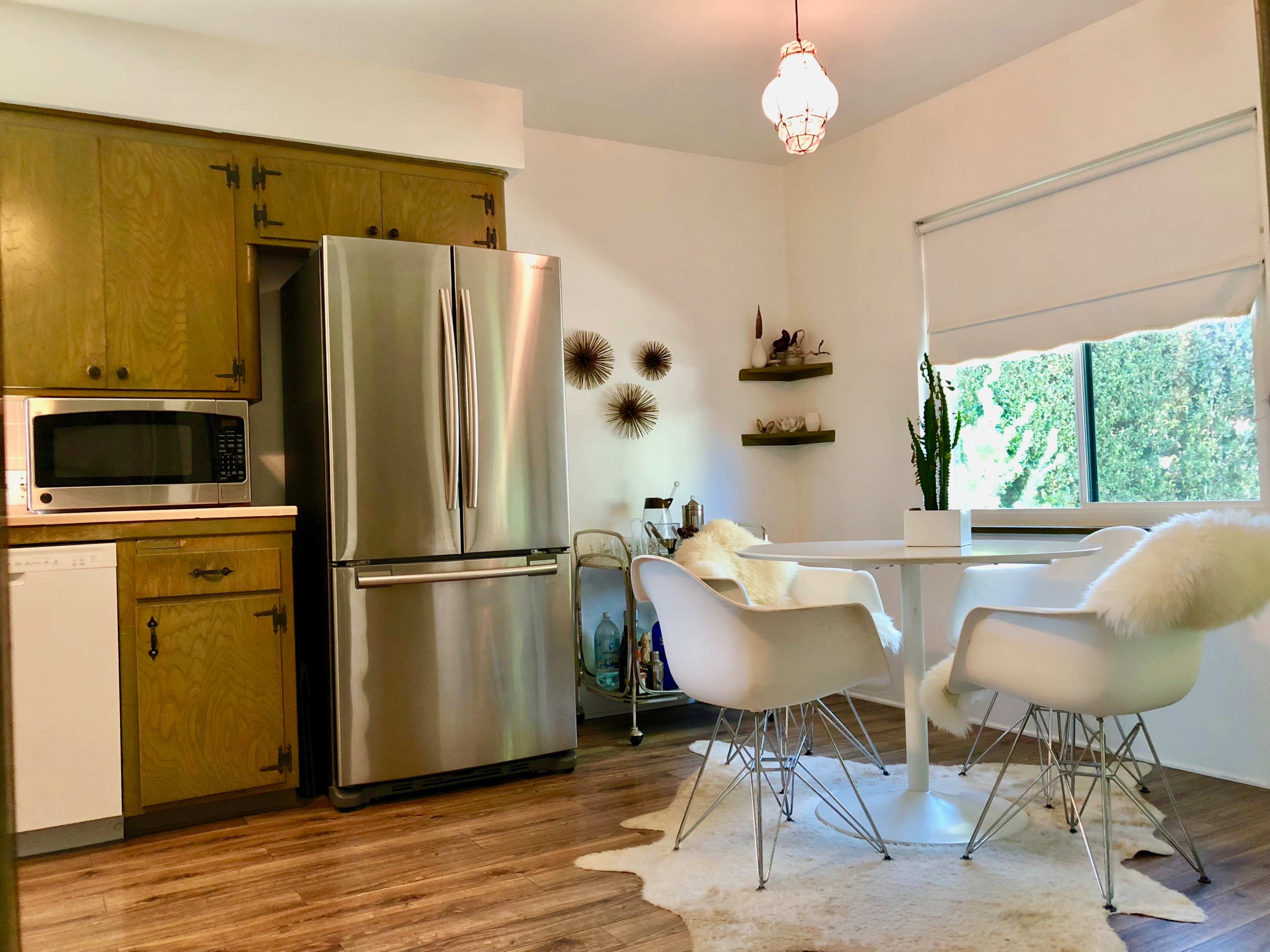 A kitchen with wooden cabinets, stainless steel appliances, and a white dining table surrounded by four chairs, with a window offering a view of greenery outside.