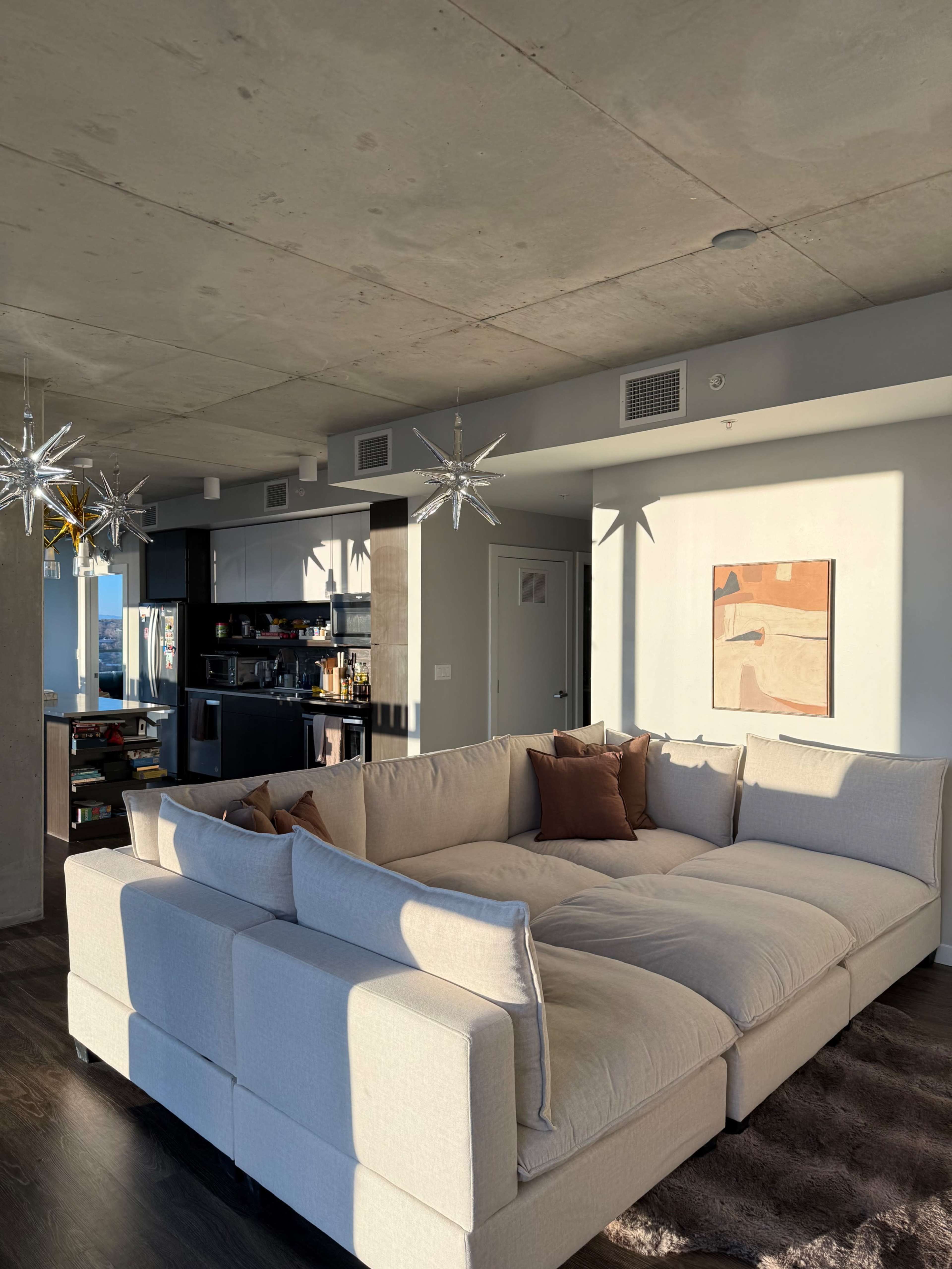 A spacious living room features a large, sectional sofa with beige upholstery and decorative pillows, illuminated by sunlight streaming through a window.