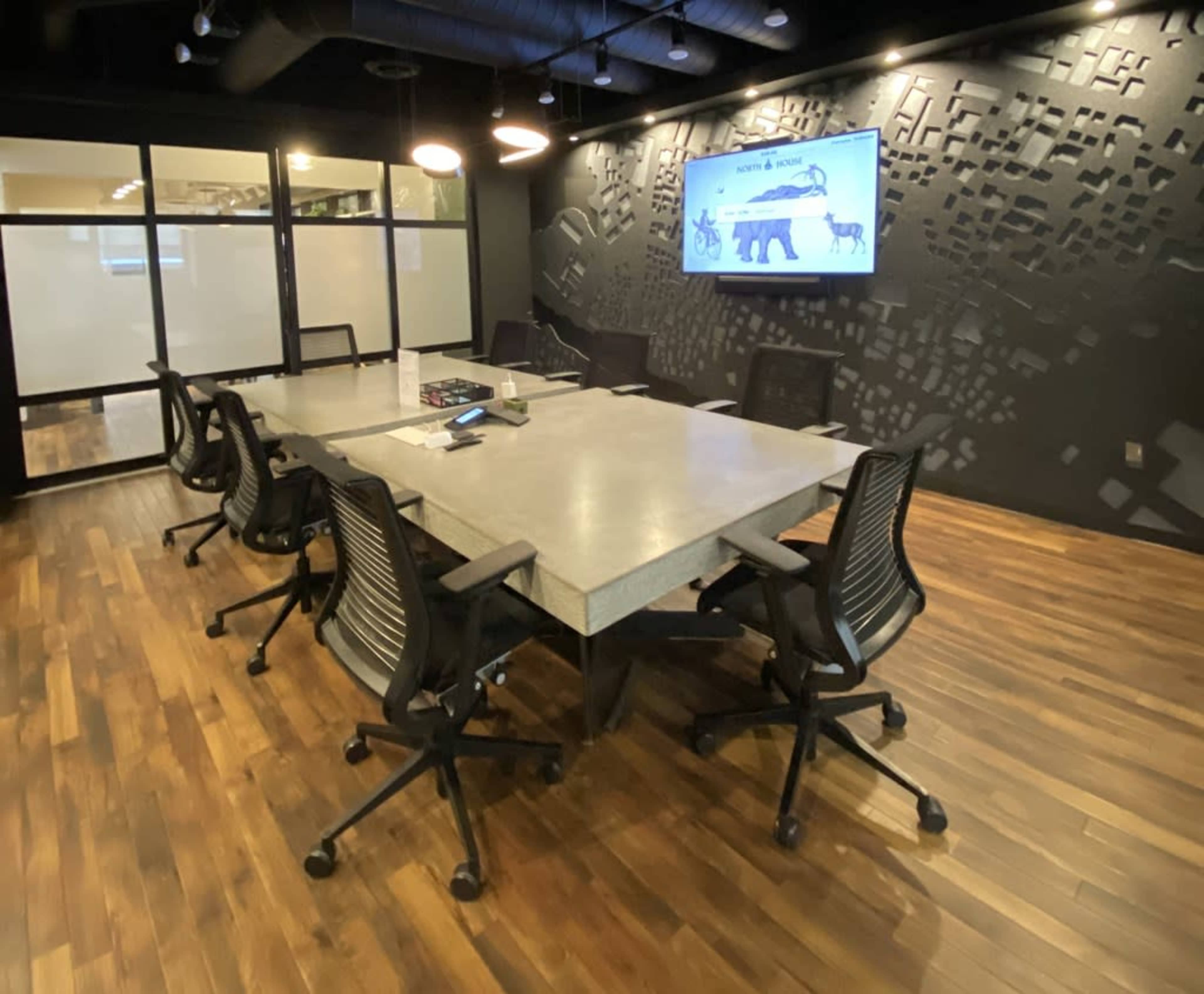 A modern conference room features a large rectangular table surrounded by black rolling chairs, with a screen displaying content on the wall and large windows partially visible.