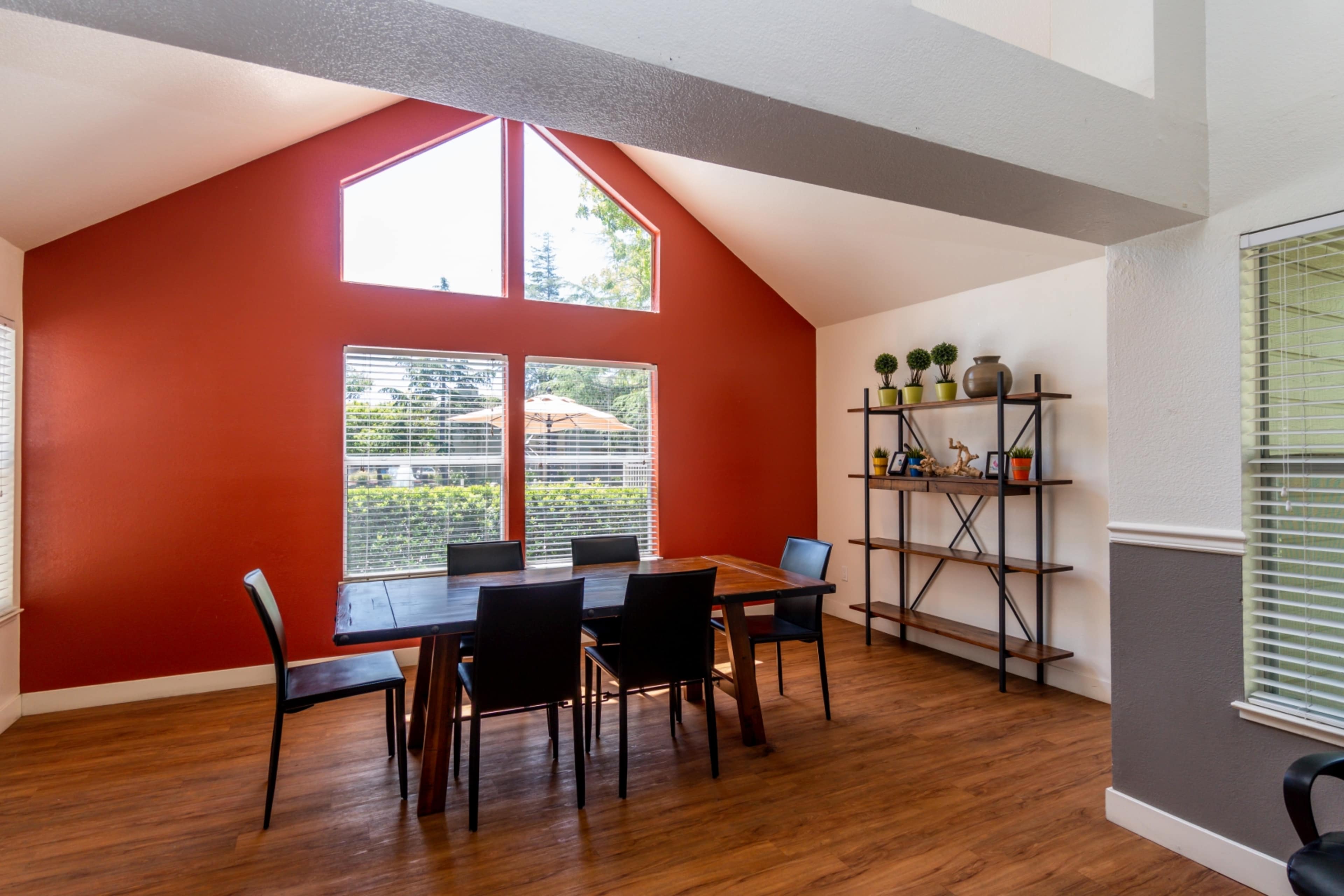 A dining area with a wooden table surrounded by black chairs, set against a red accent wall and large windows that overlook an outdoor space.