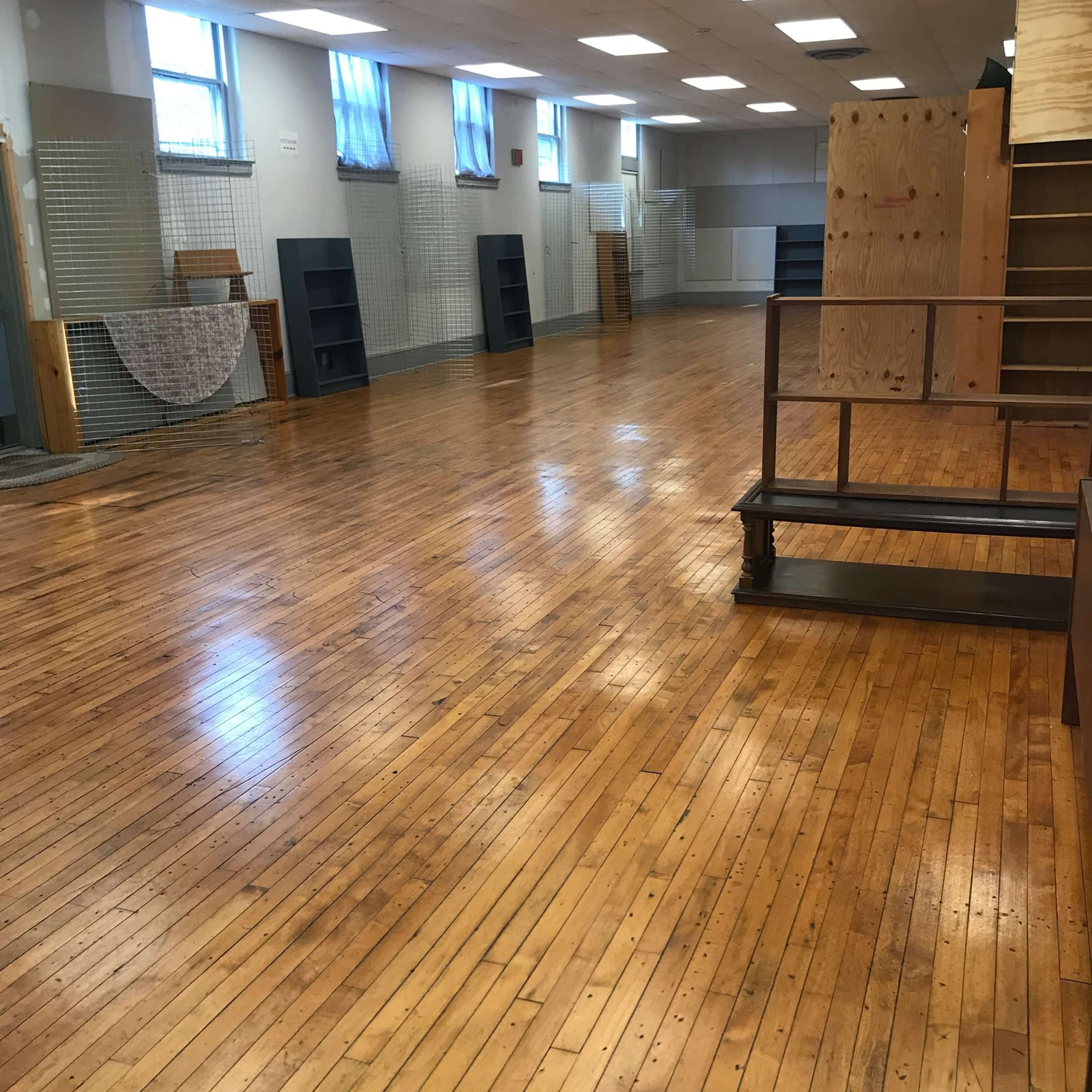 The image shows an empty, spacious room with polished wooden floors, shelves along the walls, and large windows allowing natural light to enter.