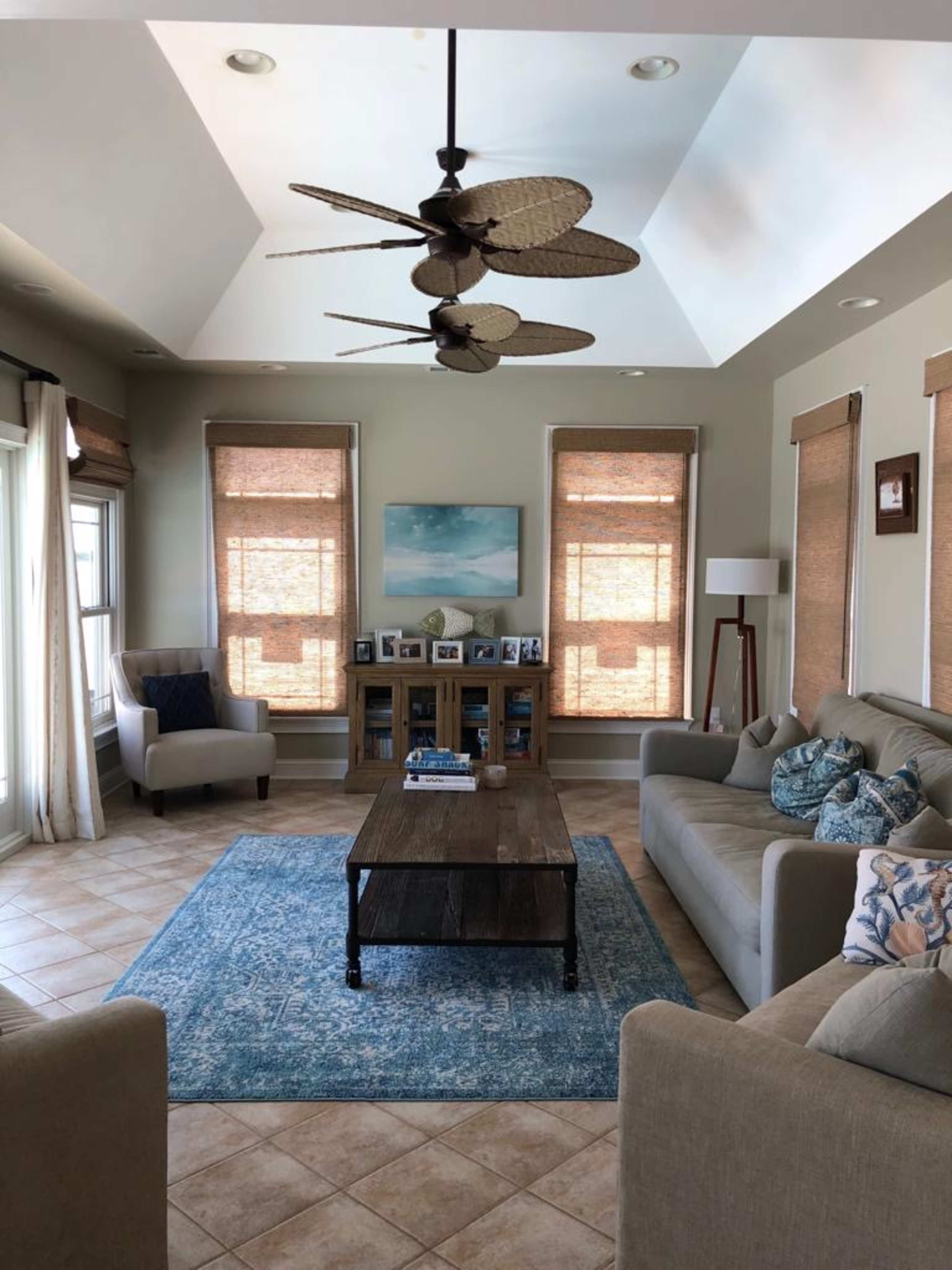 A spacious living room features a light-colored sofa, an area rug, a wooden coffee table, and large windows covered with woven shades.