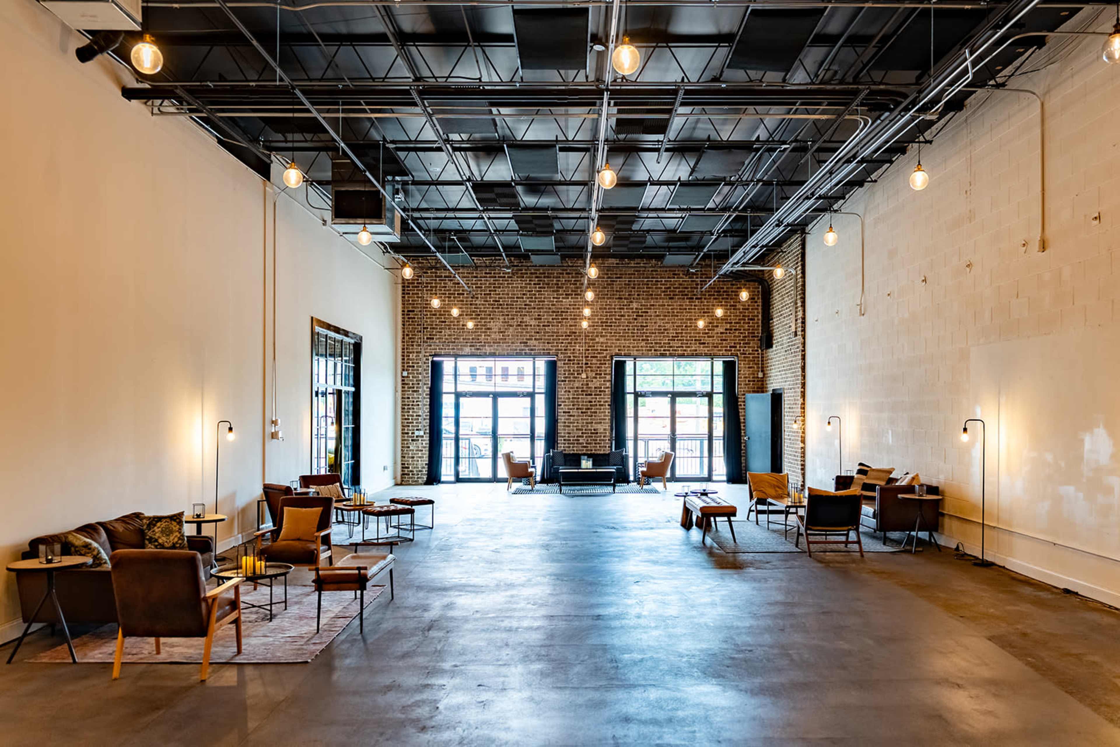 The image shows a spacious, modern interior with industrial design elements, featuring multiple seating areas and warm lighting.