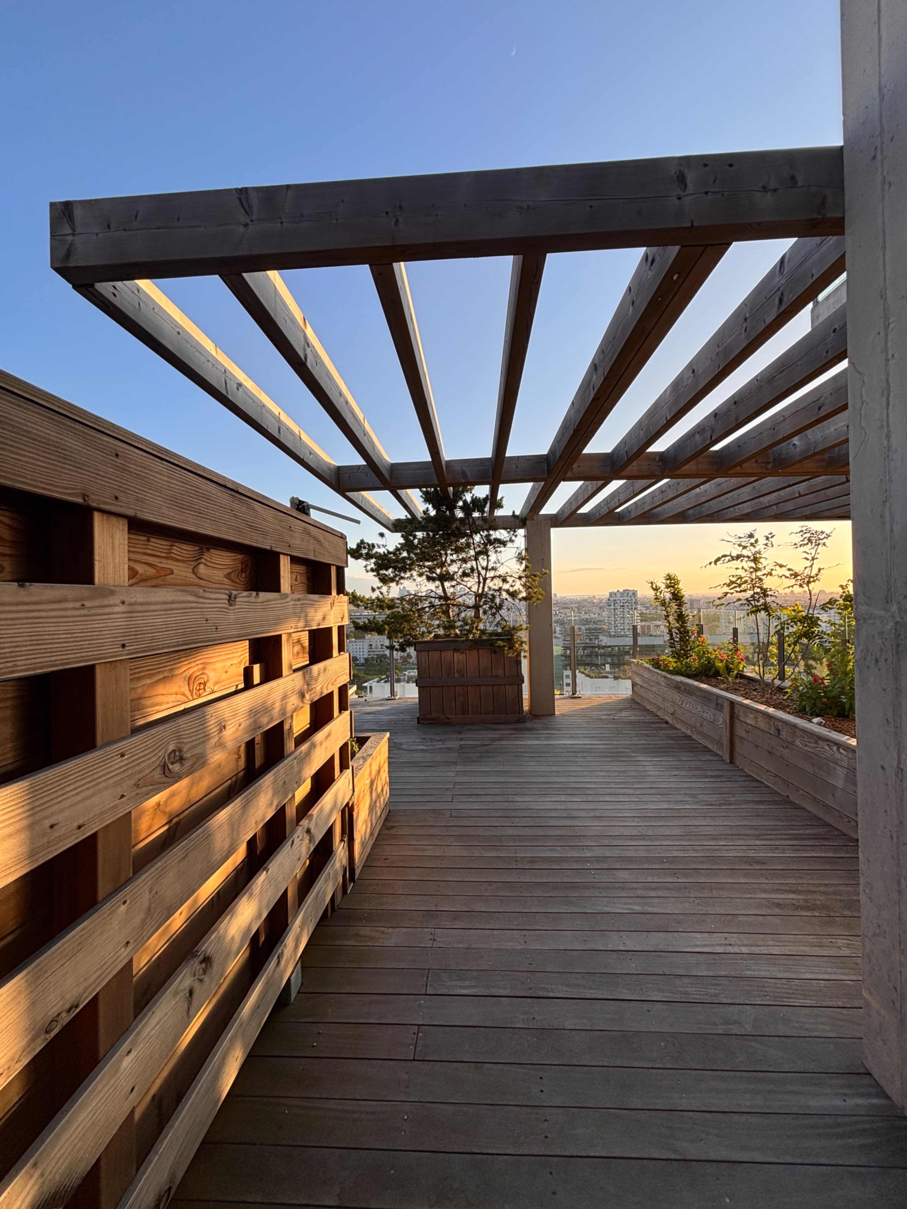 The image shows a wooden deck area with a slatted overhead structure, leading to a planter and a view of a cityscape at sunset.