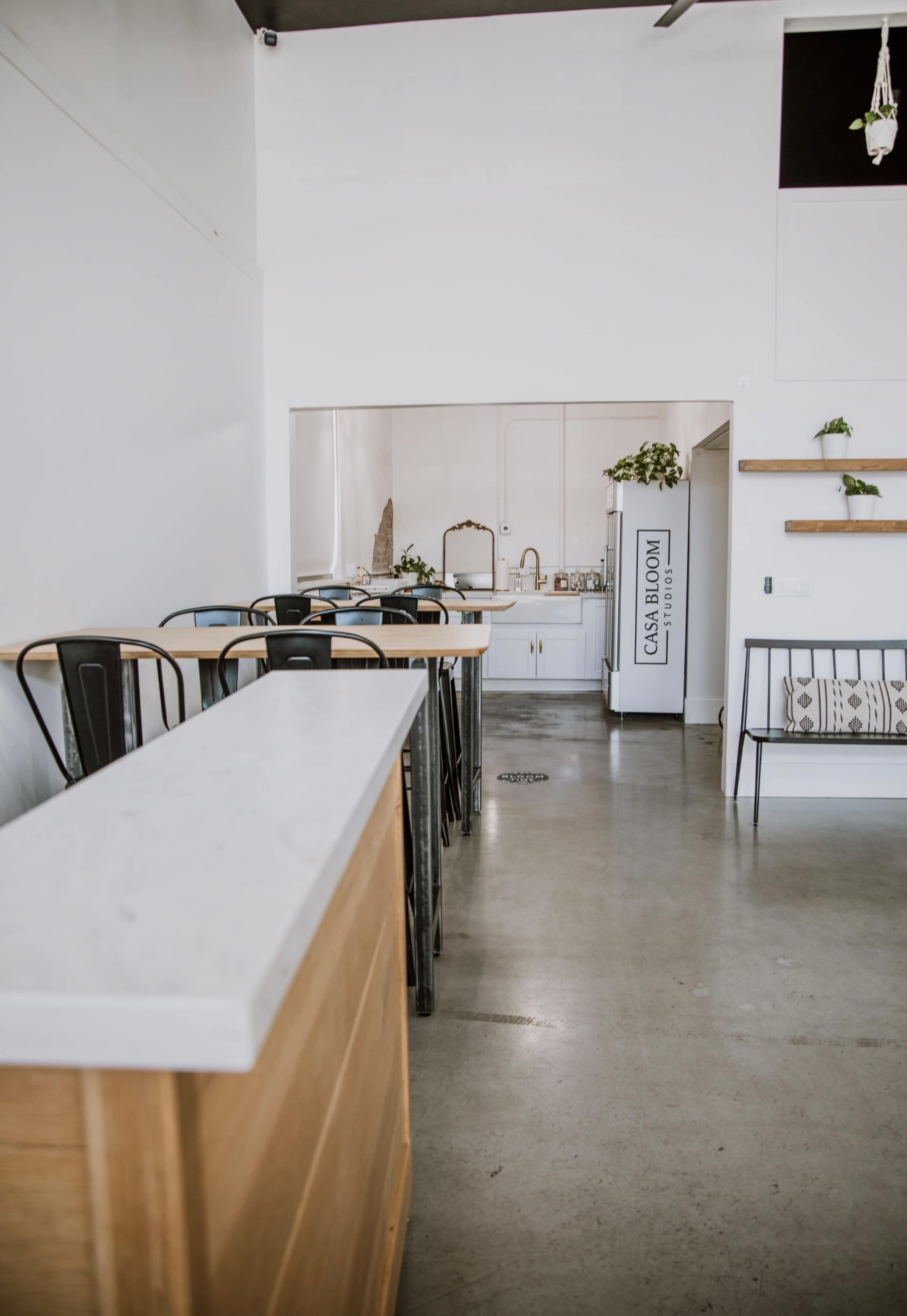 The image shows a modern, minimalistic cafe interior with a long marble countertop and several black metal chairs lining wooden tables.