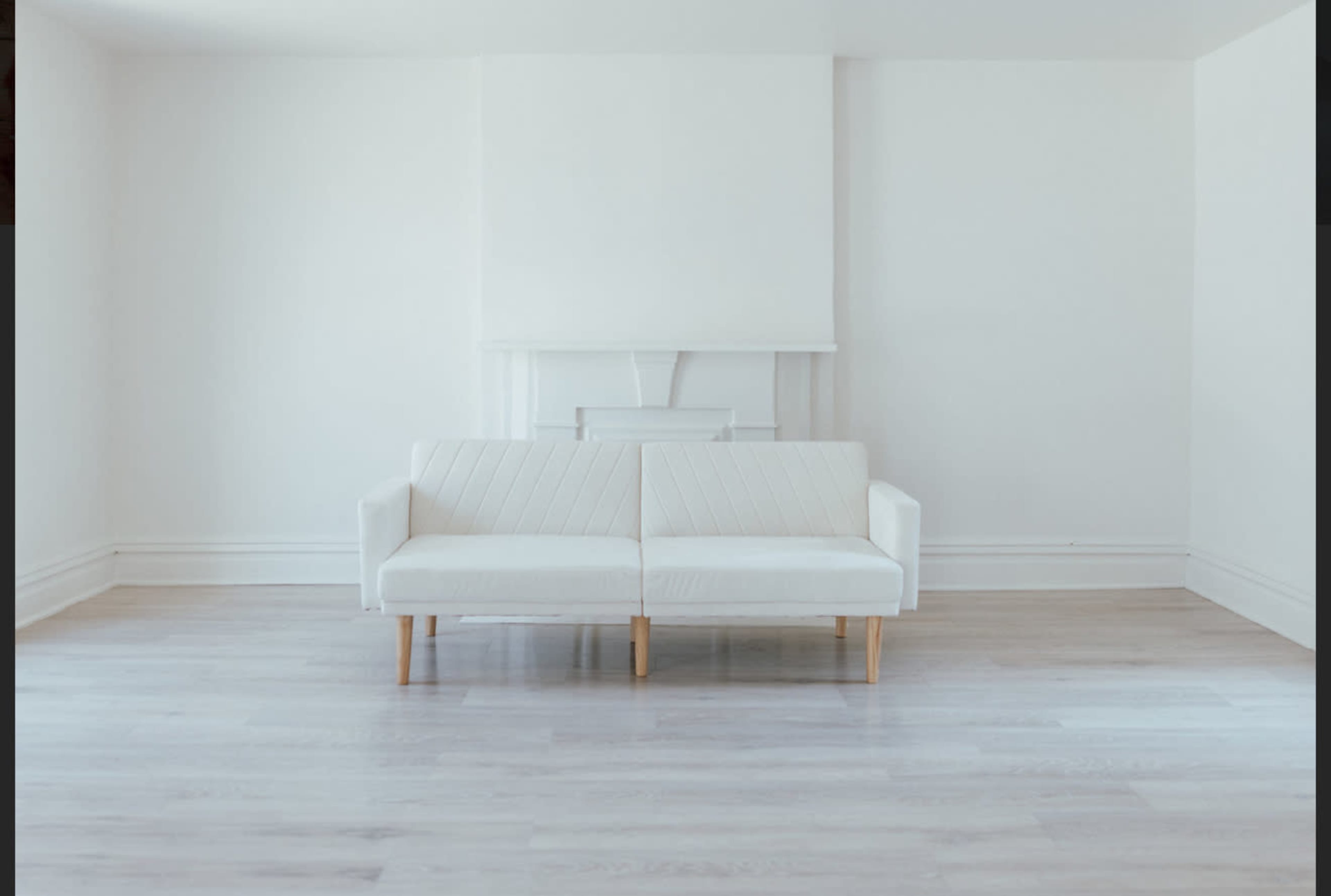 A minimalist room features a white, two-seater sofa positioned in the center, with light-colored wooden flooring and plain walls.