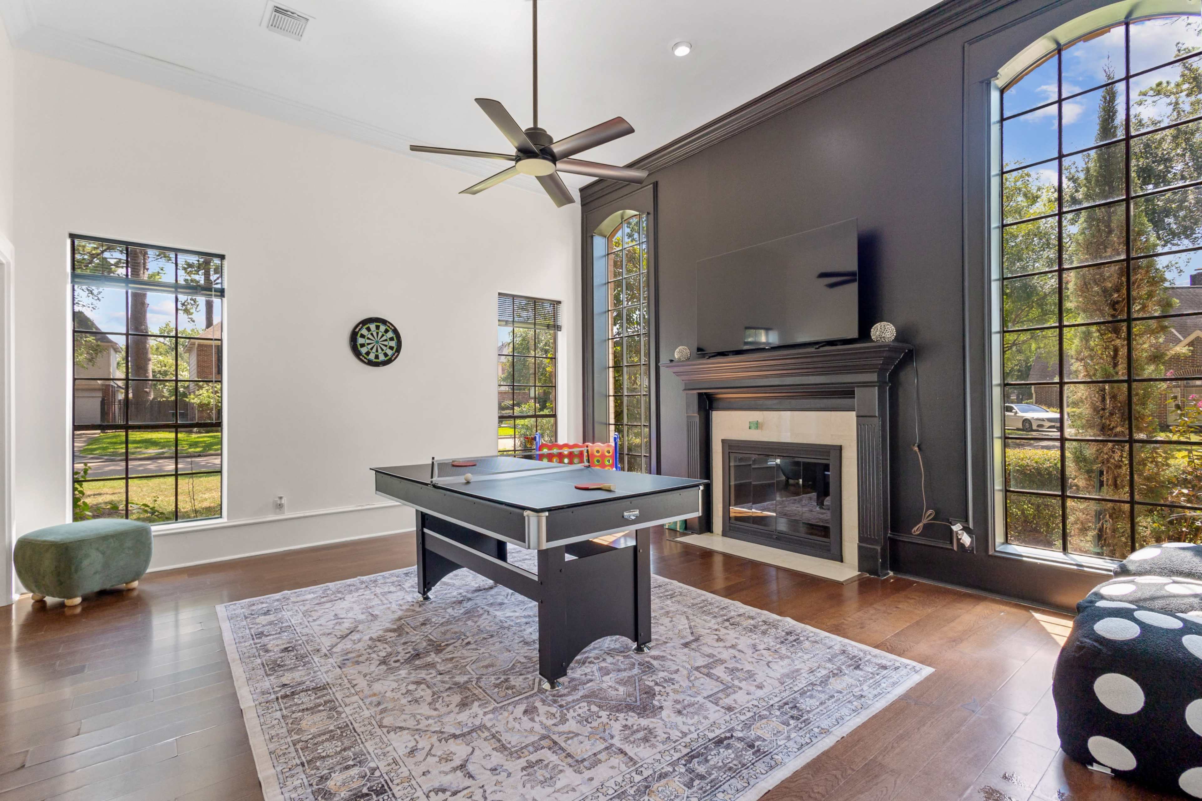 The image shows a spacious room featuring a black ping pong table, a fireplace, and large windows that let in natural light.