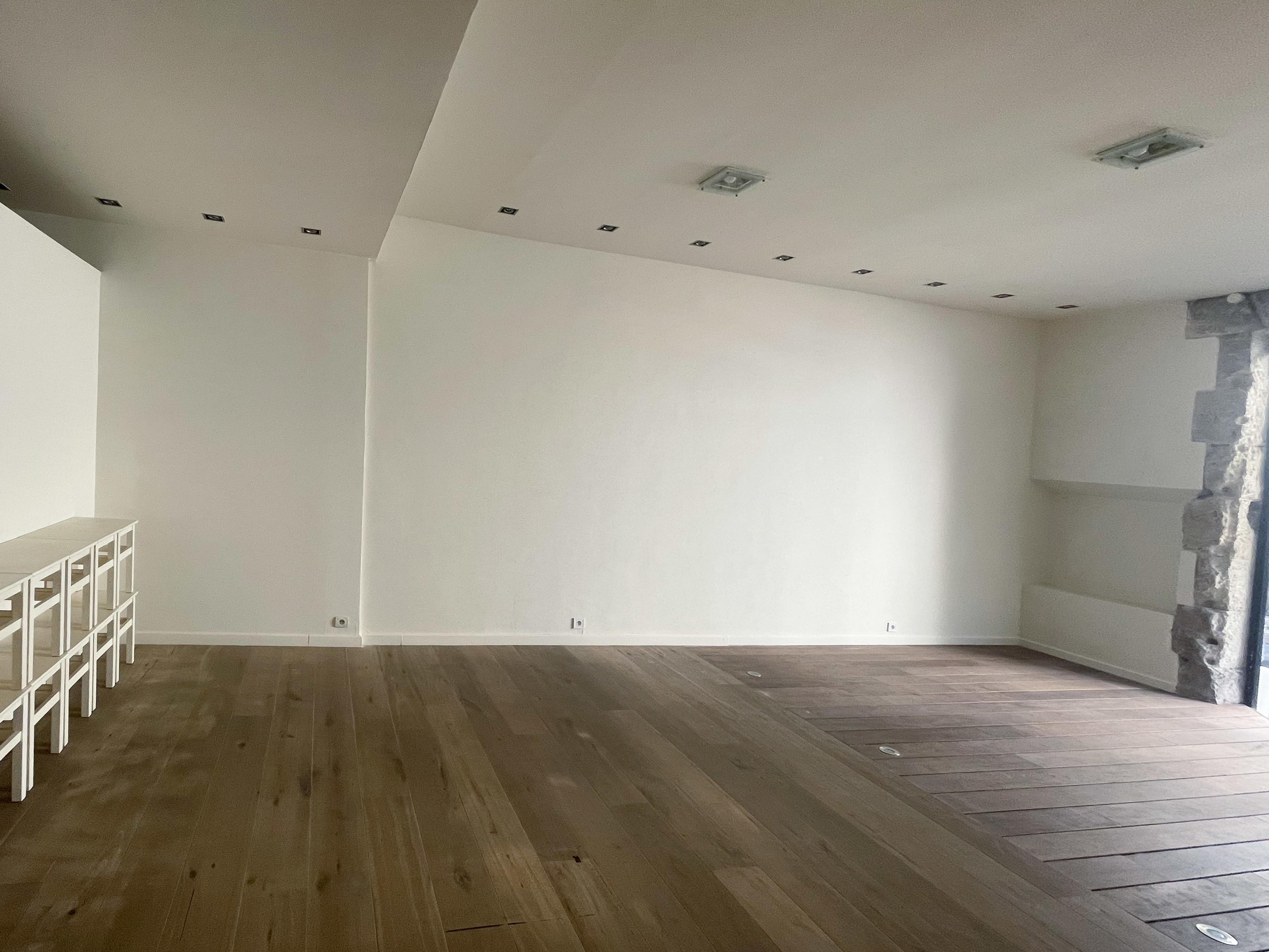 The image shows a spacious, empty room with light-colored walls, wooden floors, and recessed lighting.