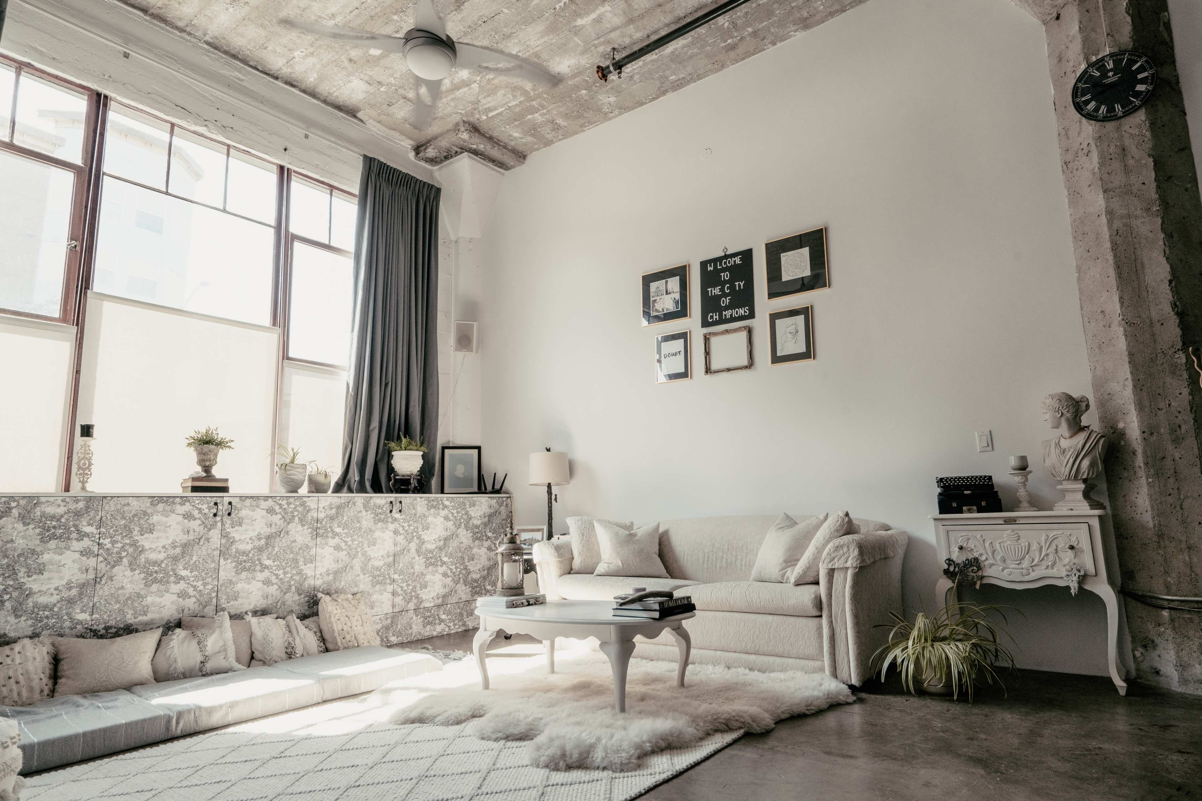 The image shows a modern living room with a light-colored sofa, a coffee table, and large windows allowing natural light to enter the space.