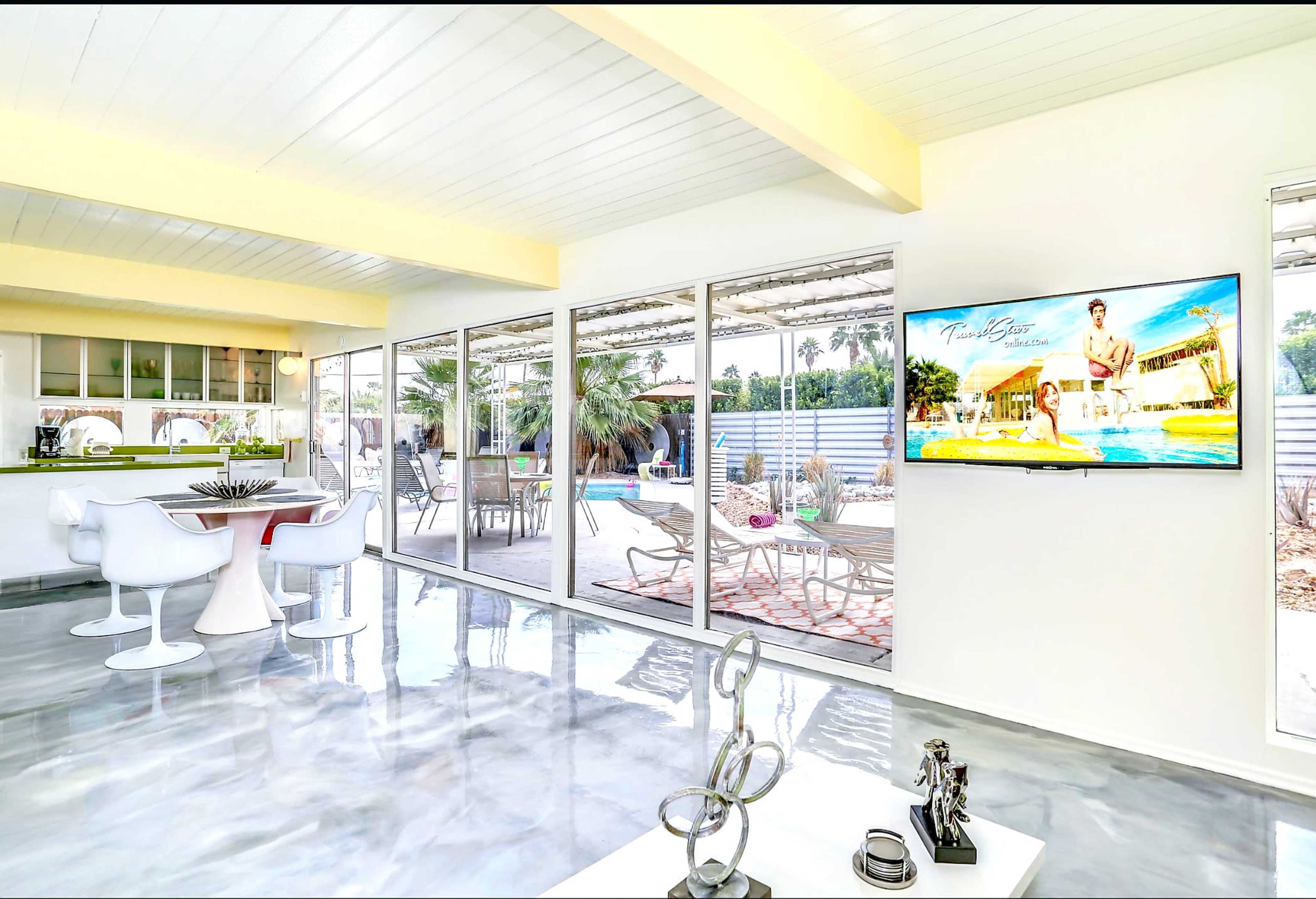 The image shows a modern, open-concept living space with a view of a patio and pool area, featuring a sleek kitchen, a dining area, and a television mounted on the wall.