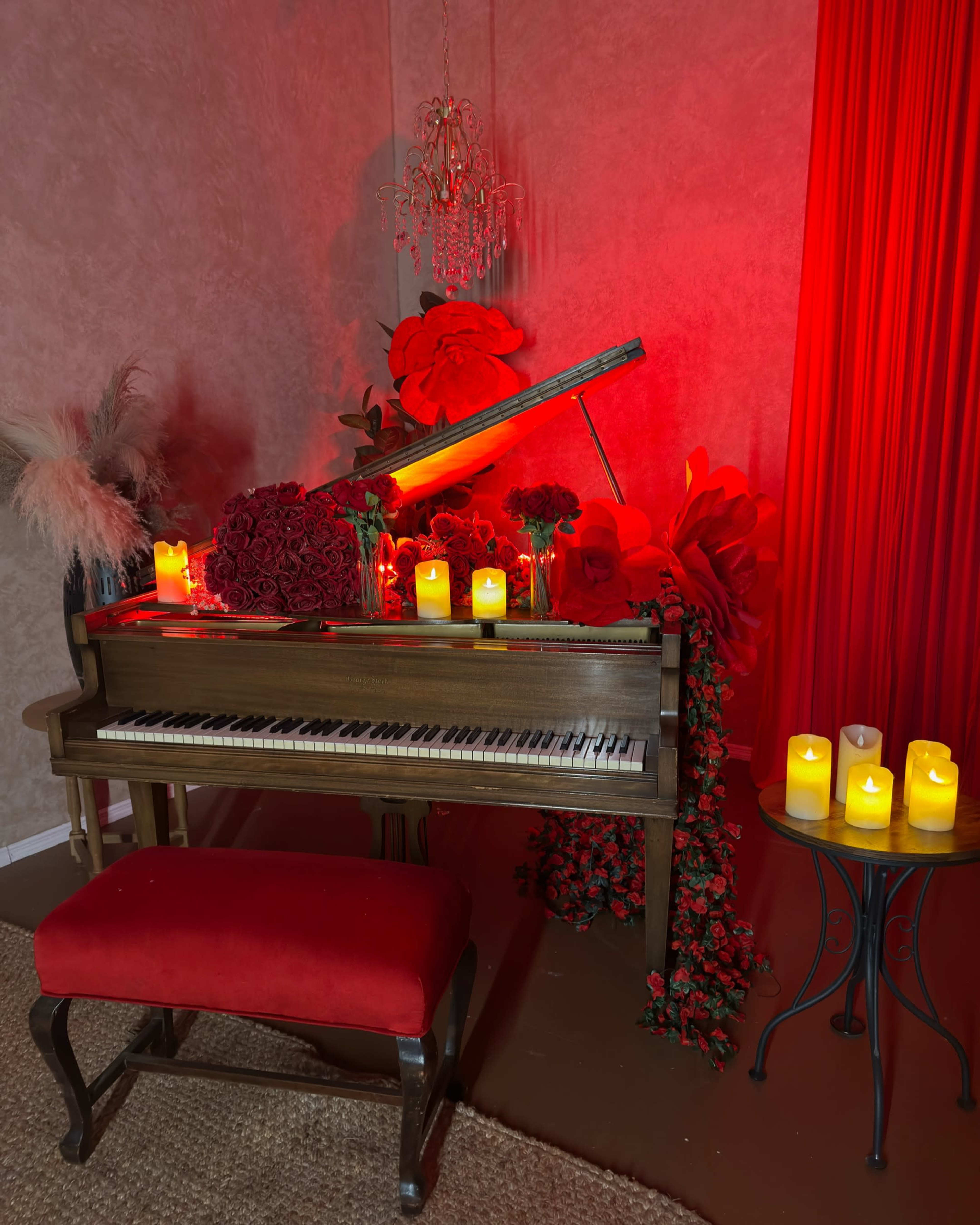 A grand piano adorned with red roses and decorative candles is positioned against a backdrop of red fabric and floral arrangements.
