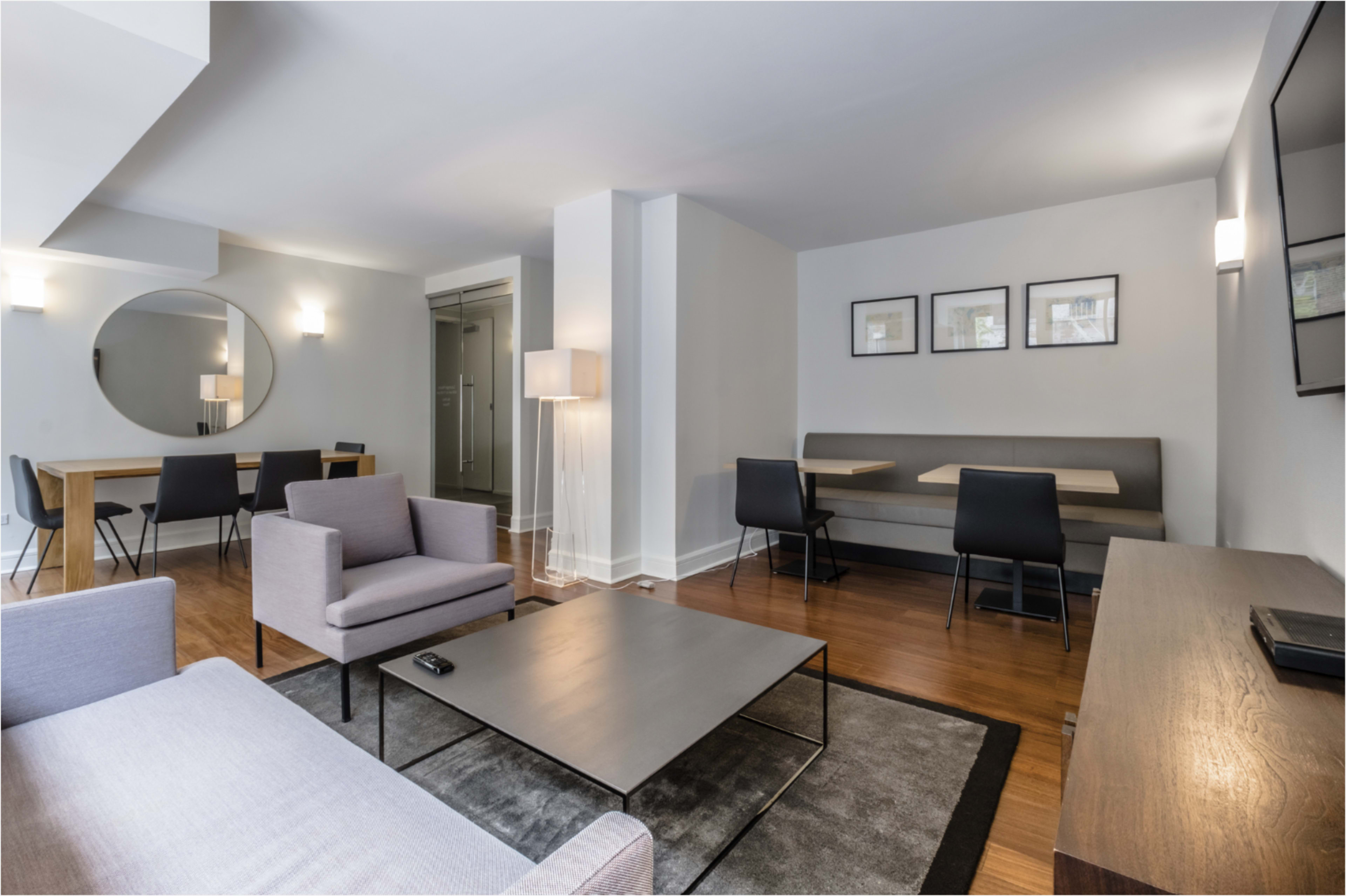 The image shows a modern living area featuring two gray armchairs, a black coffee table, a dining table, a wall-mounted lamp, and a large round mirror.