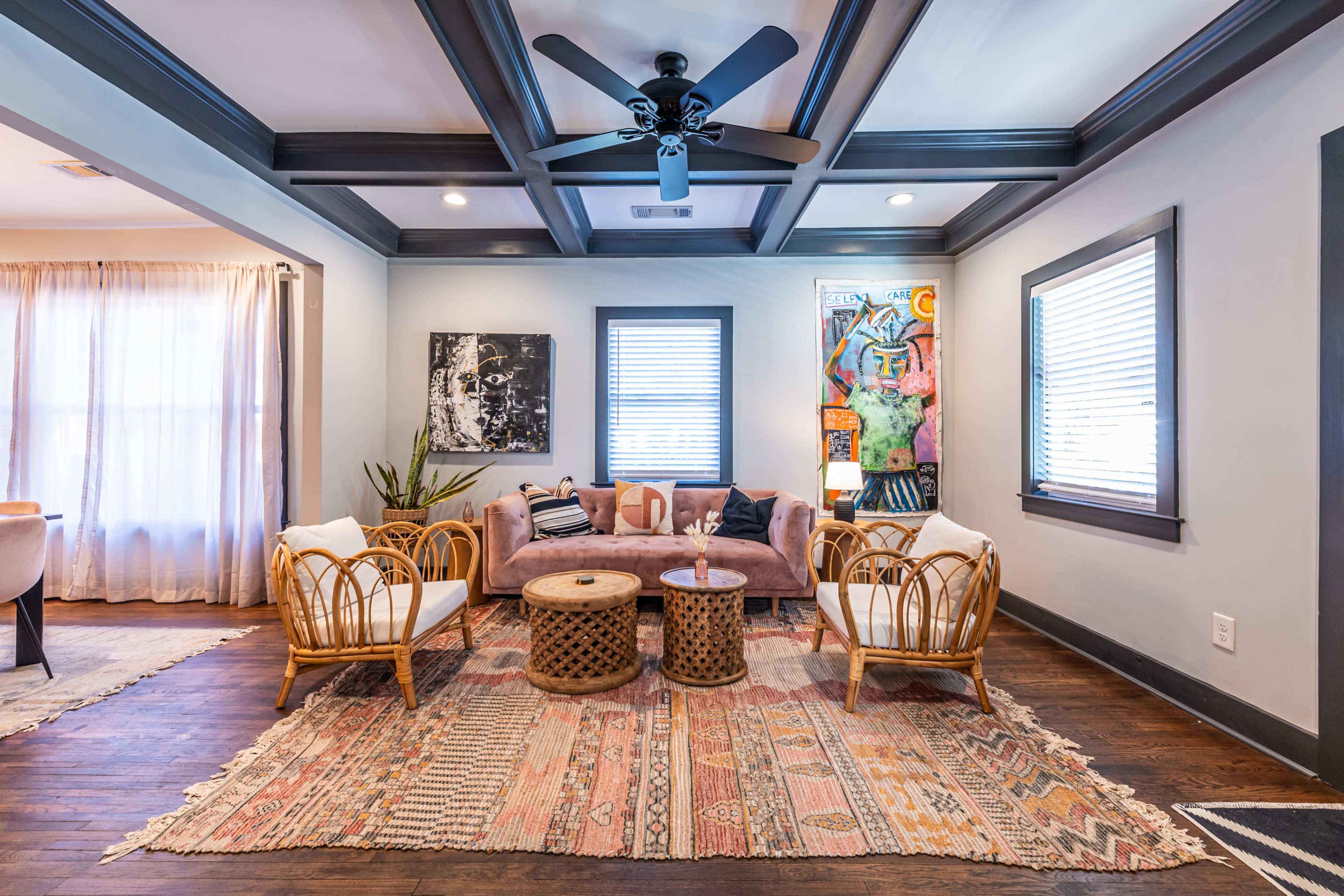 The image shows a modern living room with a coffered ceiling, featuring two rattan chairs, a pink sofa, a round wooden coffee table, and colorful artwork on the walls.