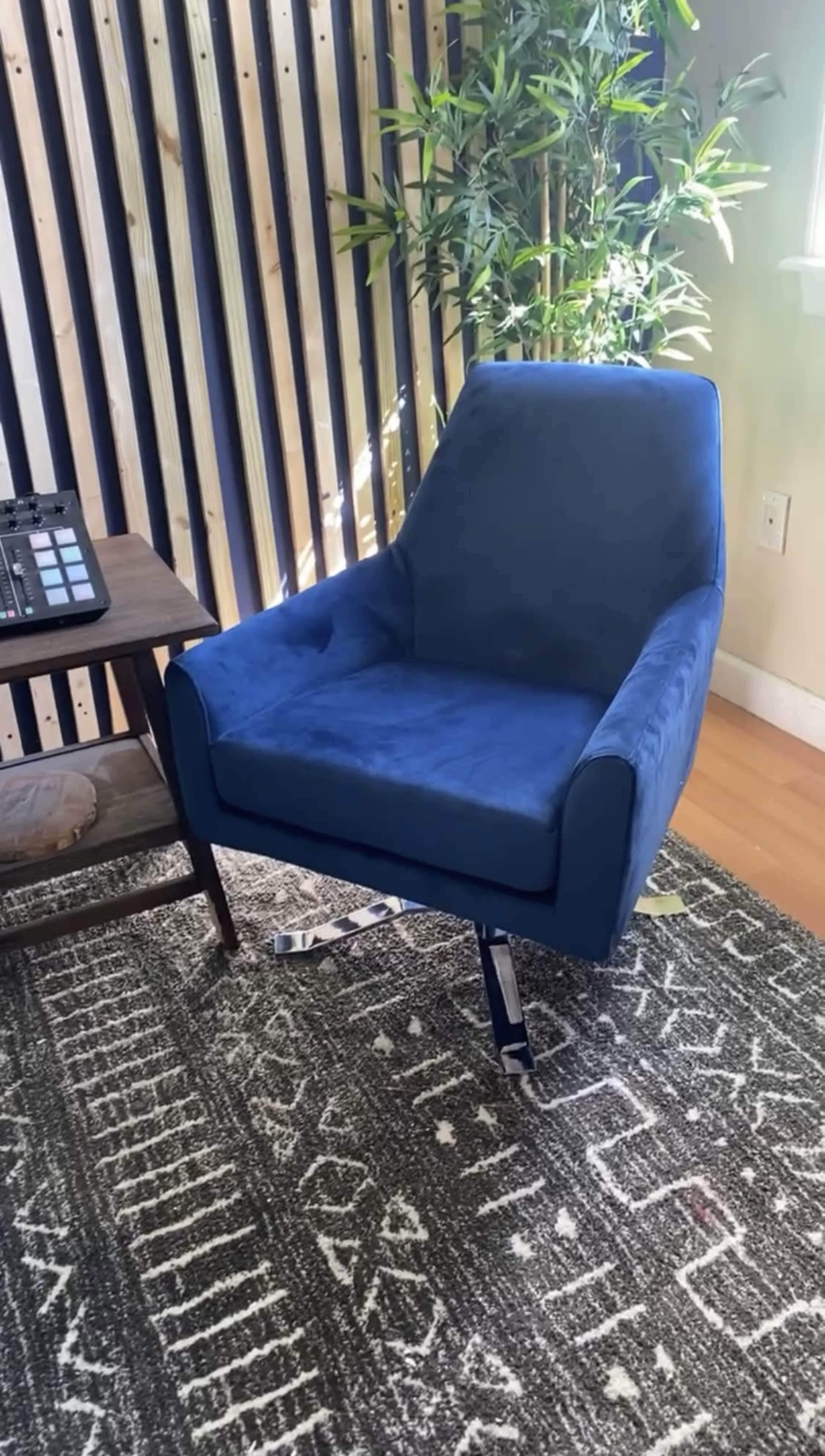 The image shows a blue upholstered chair positioned next to a wooden table with an electronic device and a plant in a room with patterned flooring and vertical wooden wall slats.