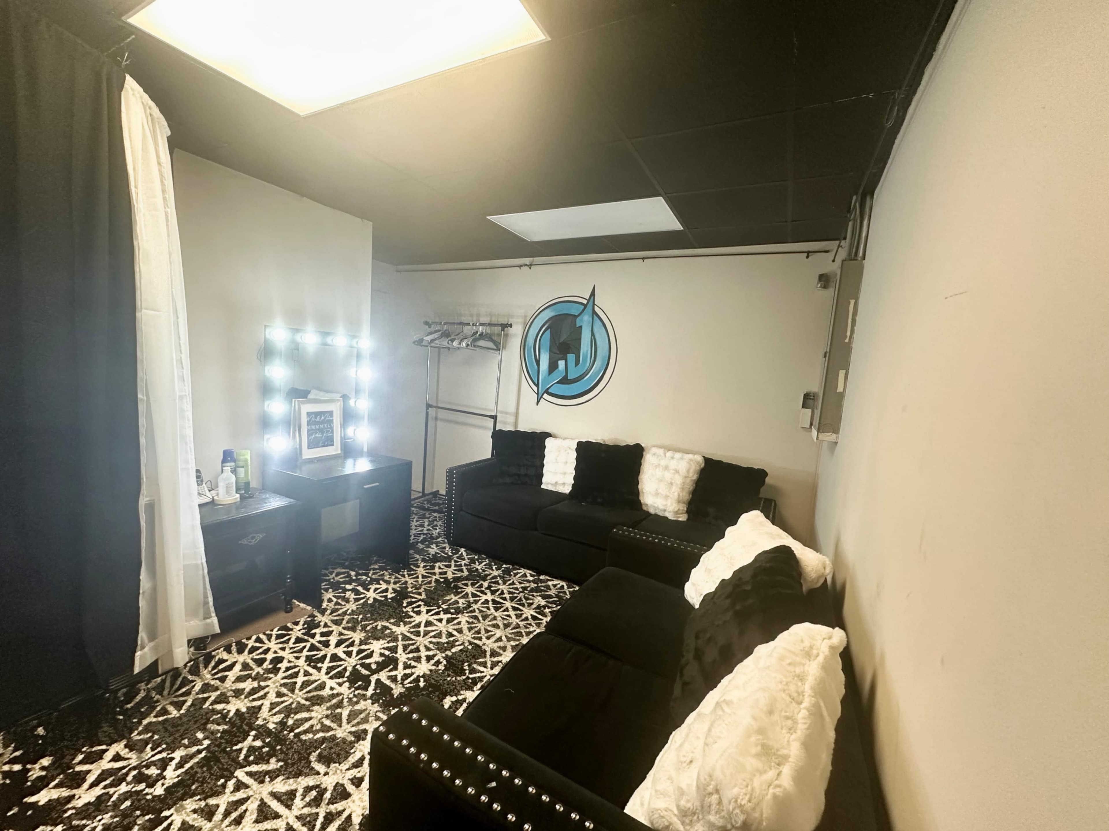 A small, modern lounge area with a black and white patterned carpet, a black sectional sofa, a vanity mirror with lights, and a wall featuring a stylized logo.