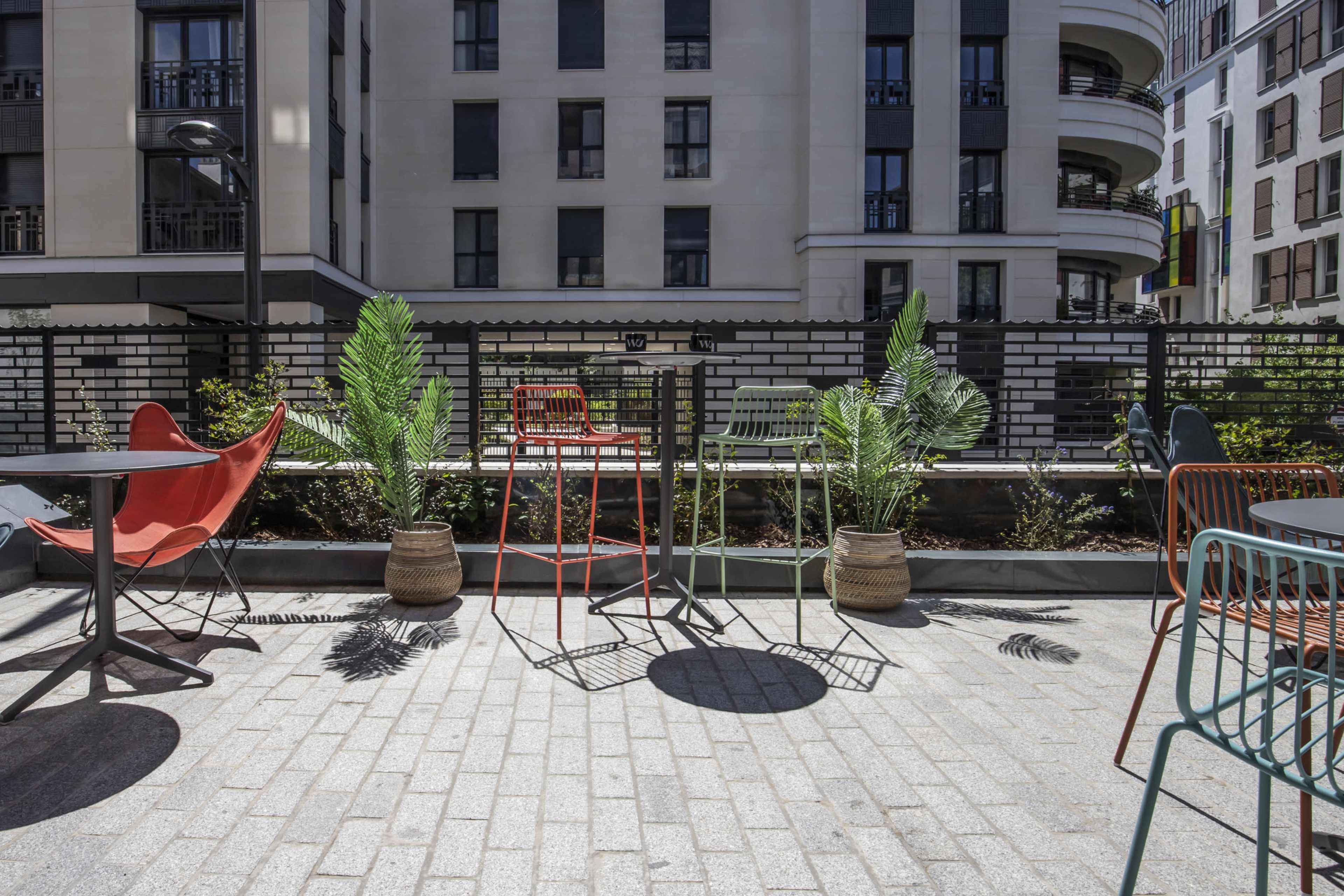 The scene features a modern outdoor seating area with colorful chairs, potted plants, and a paved terrace surrounded by a contemporary building.