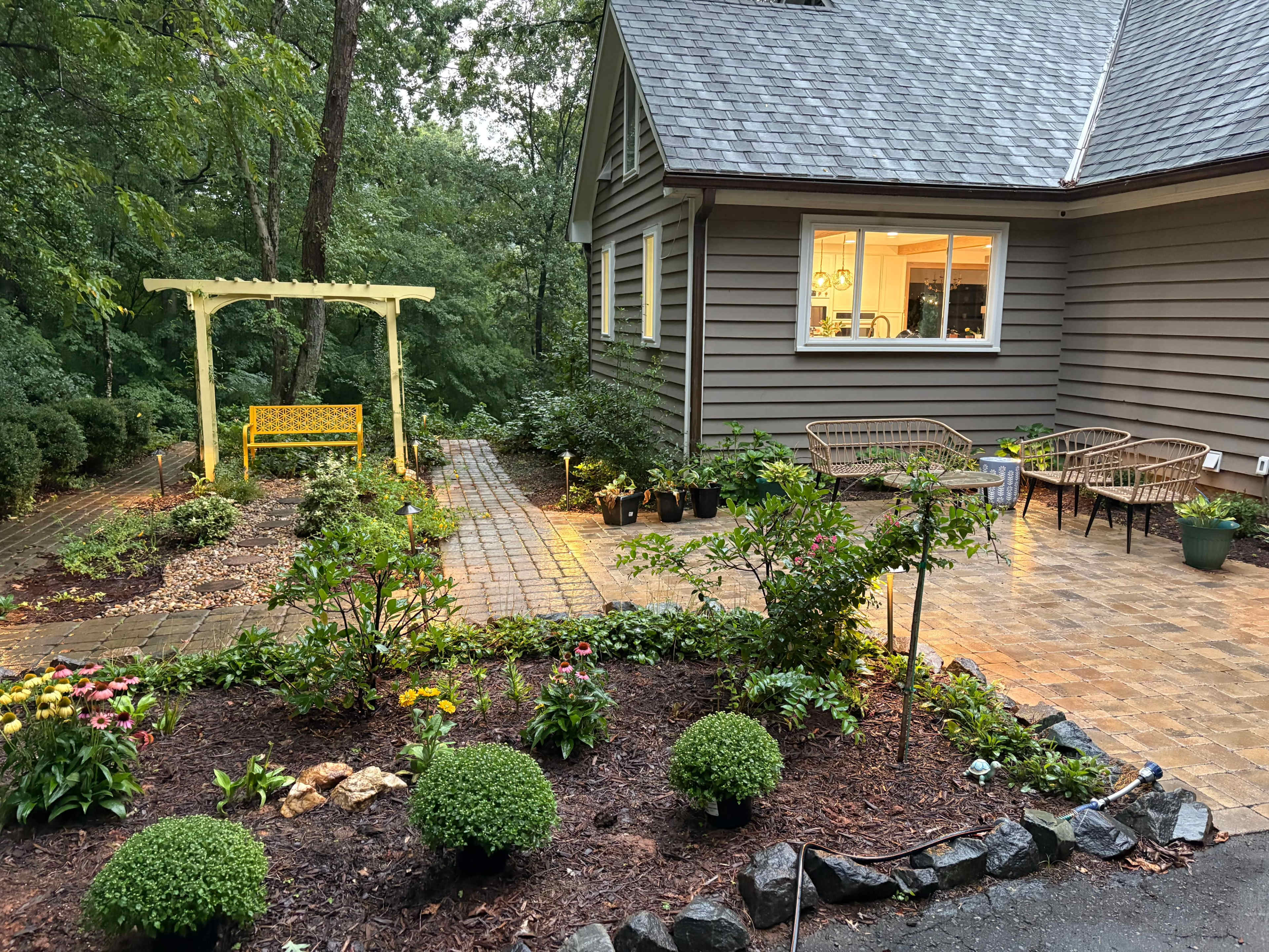 The image shows a landscaped backyard with a yellow bench beside a stone pathway, surrounded by various plants and seating areas near a house.