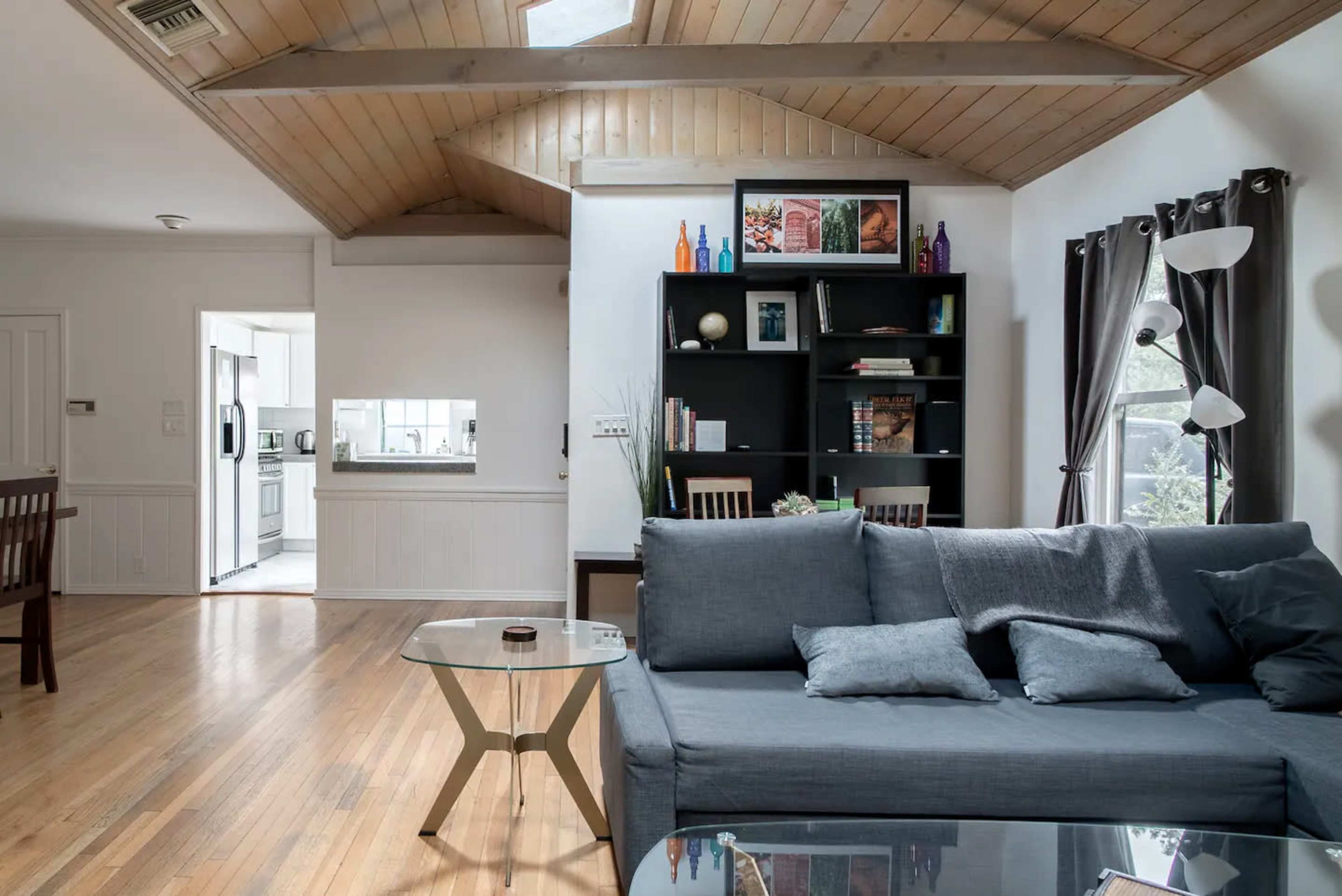 The image shows a modern living room featuring a gray sofa, a glass coffee table, and a black bookshelf, with visible access to a kitchen in the background.