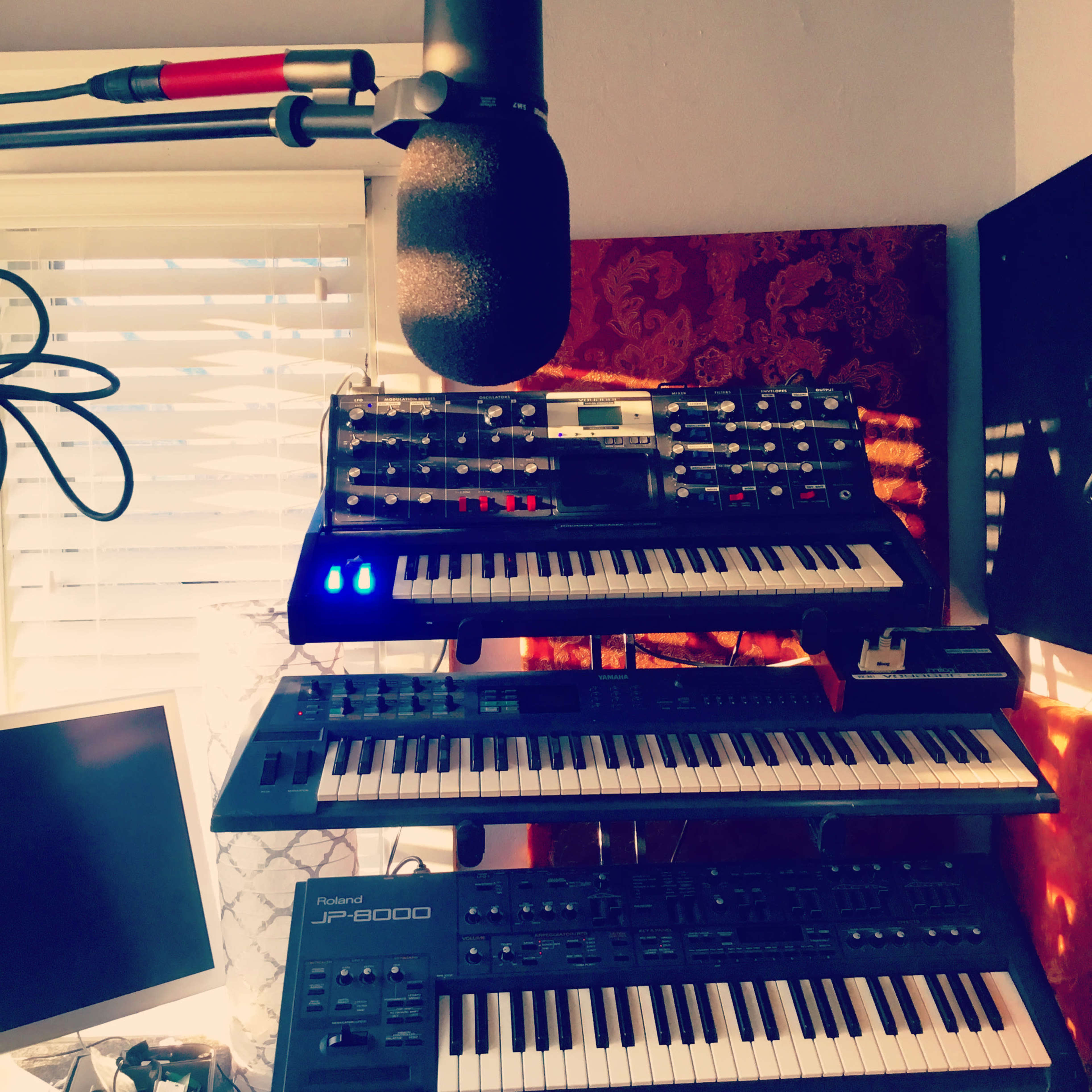 A music production setup featuring a microphone, multiple synthesizers, and a computer monitor against a backdrop of window blinds.