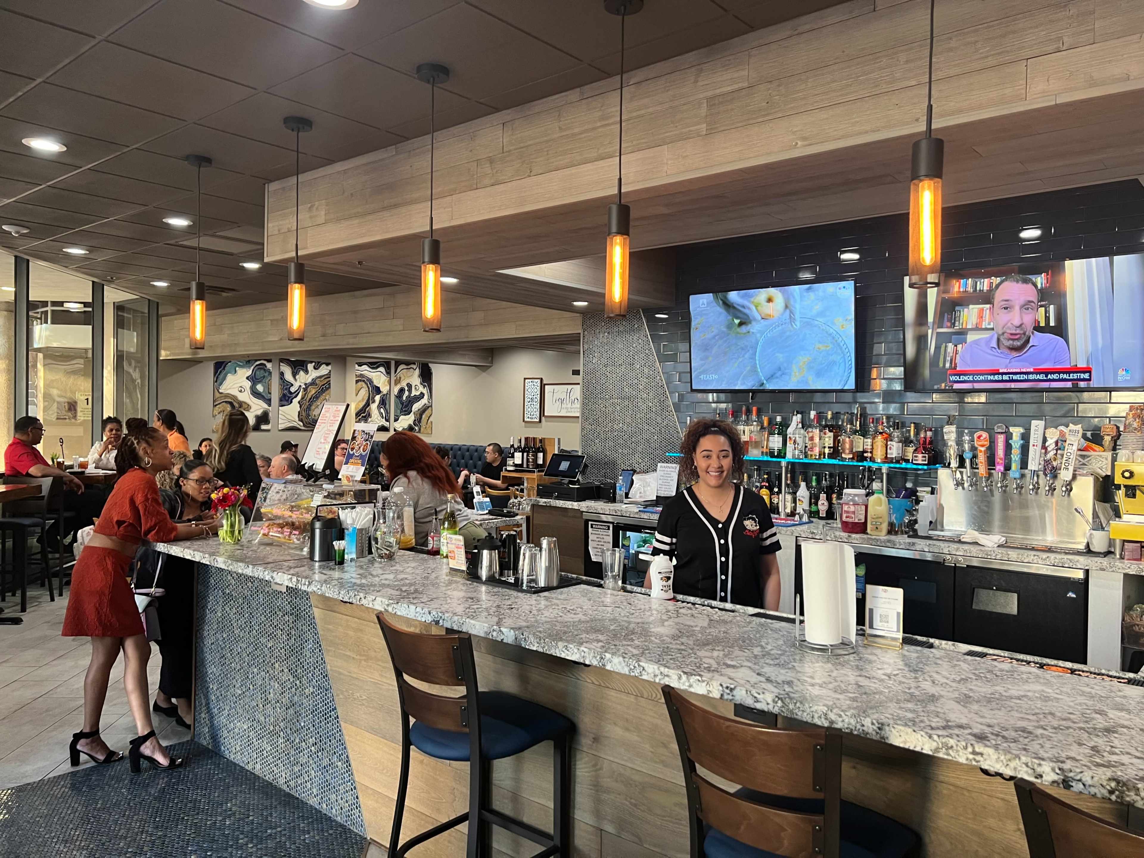 A busy bar with patrons sitting at the counter and enjoying drinks, while a bartender prepares beverages in front of screens displaying a news broadcast.