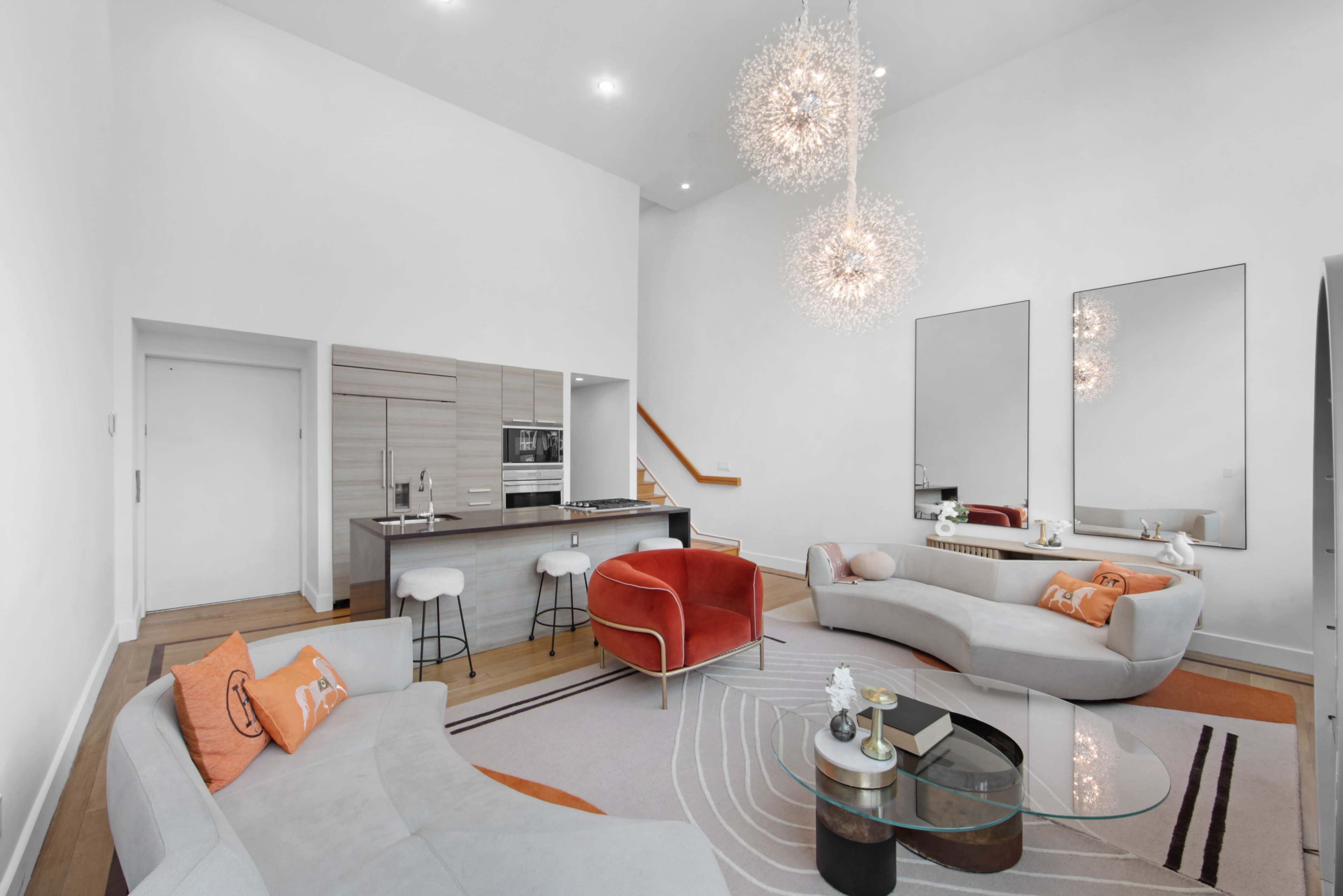 The image shows a modern living room with two curved sofas, a glass coffee table, and a kitchen area featuring bar stools and sleek cabinetry.