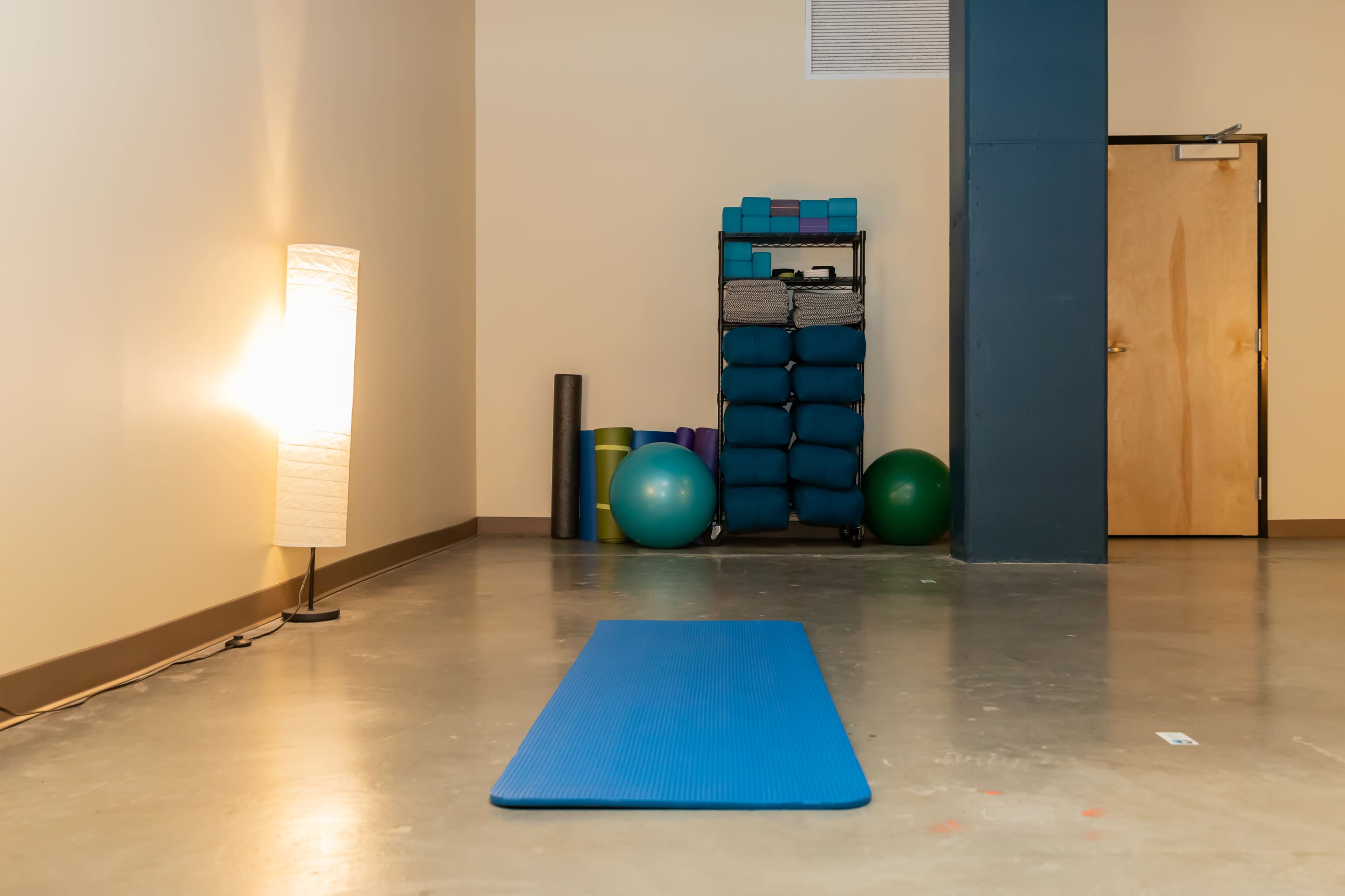 A blue yoga mat on a concrete floor in a room with exercise equipment, including yoga blocks and exercise balls stacked on a cart, and a lamp providing light.