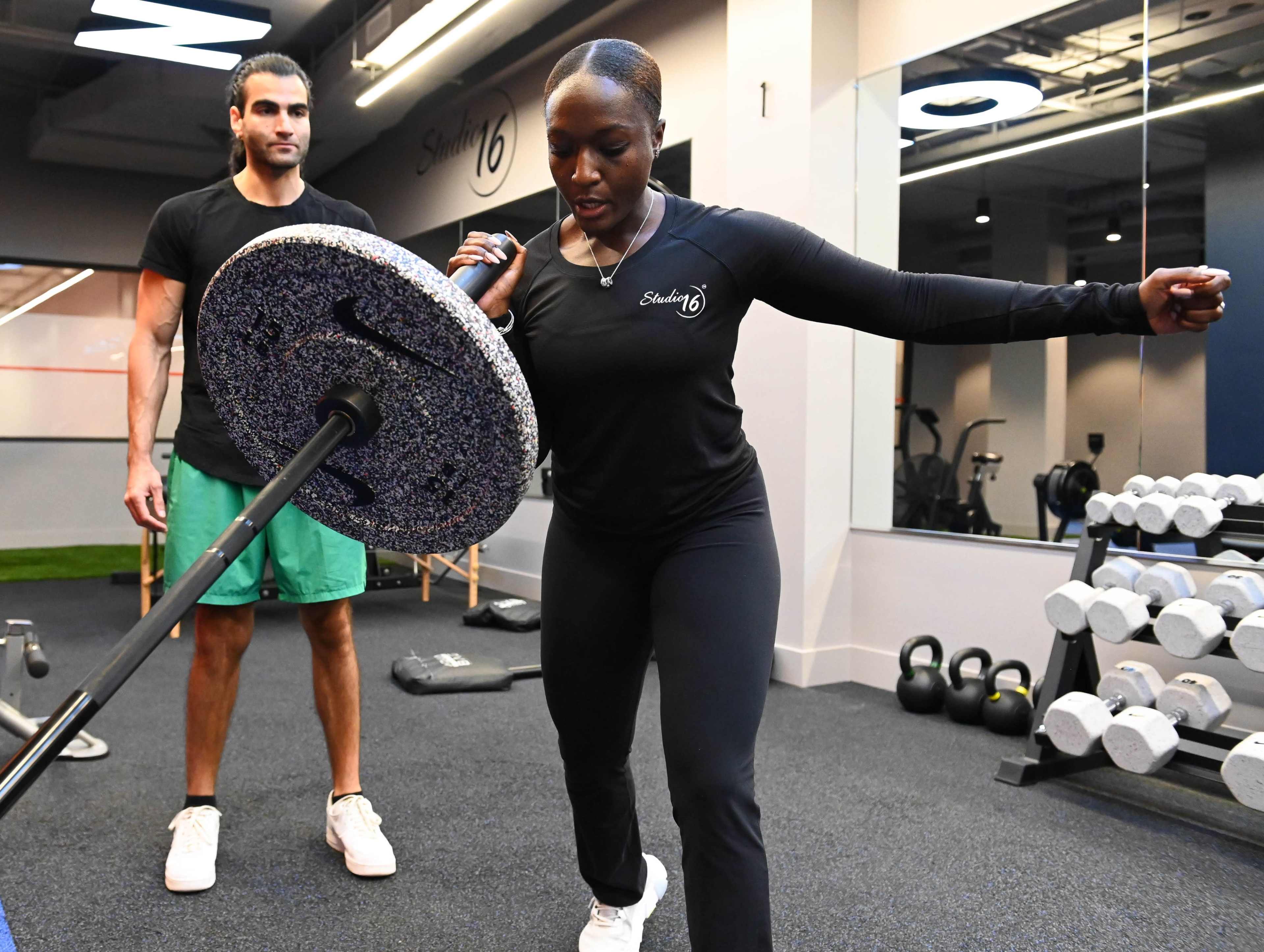 A woman exercises with a weighted barbell while a man observes and provides guidance in a gym setting.