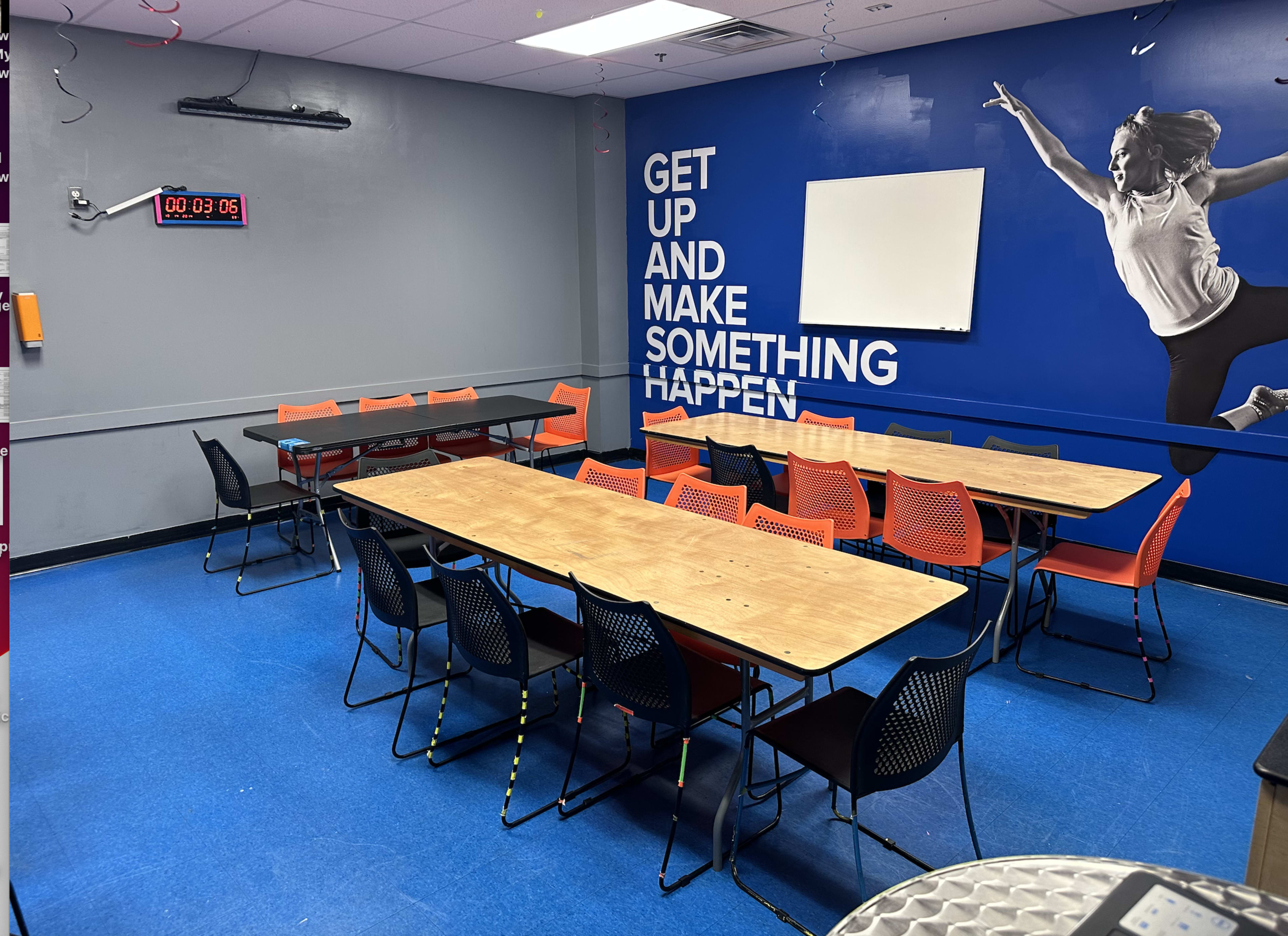 A conference room is shown with several tables arranged in a U-shape, surrounded by black and orange chairs, and a blue wall featuring a motivational phrase and a large empty whiteboard.
