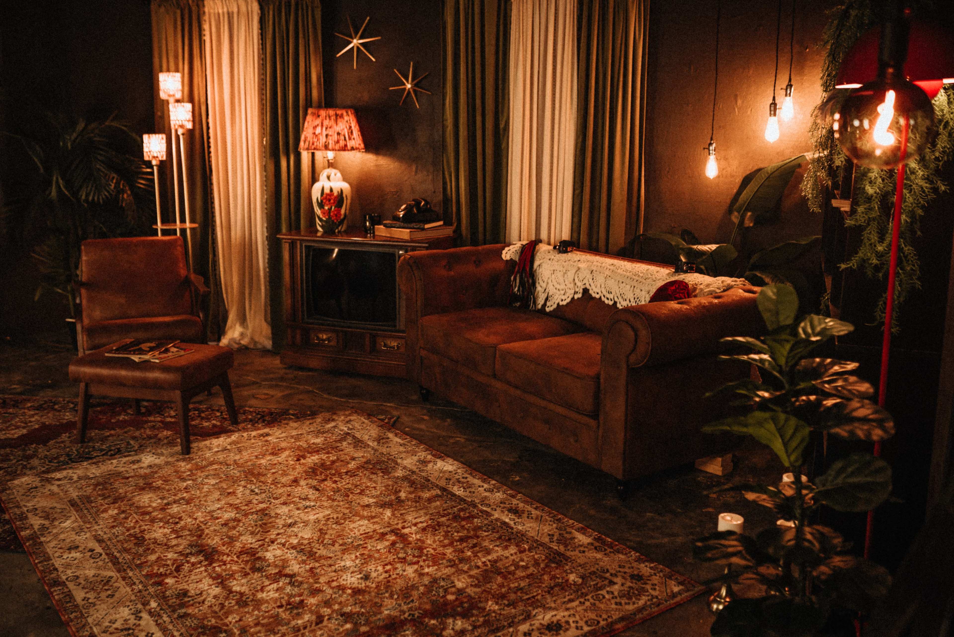 The image depicts a cozy living room with a brown sofa, a single chair, a patterned rug, and various lighting fixtures illuminating the space.