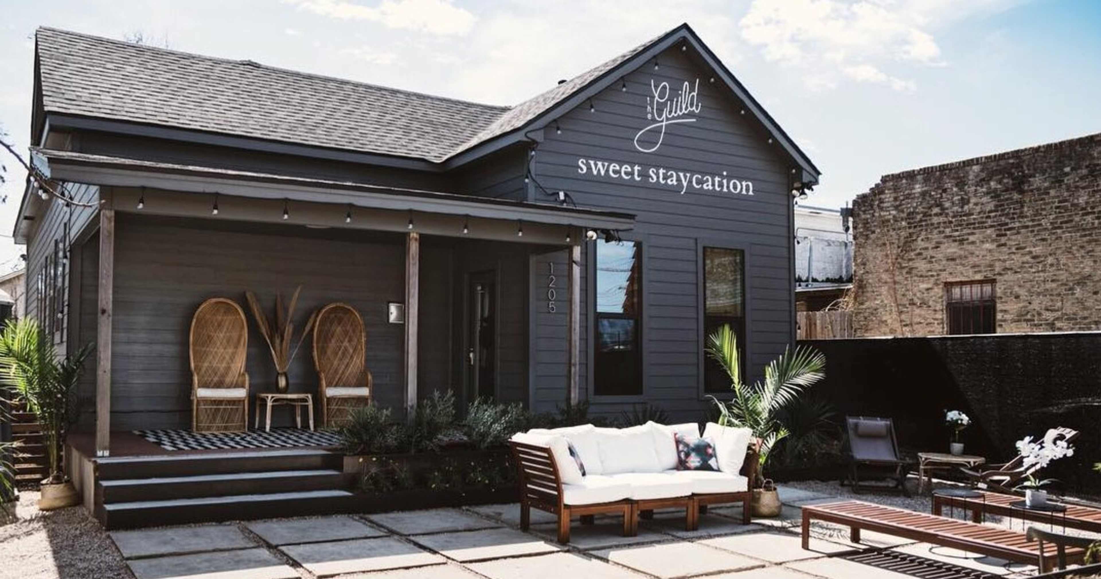 A gray house with a sign reading "sweet staycation" features a front porch with wicker chairs and a stone-paved patio.
