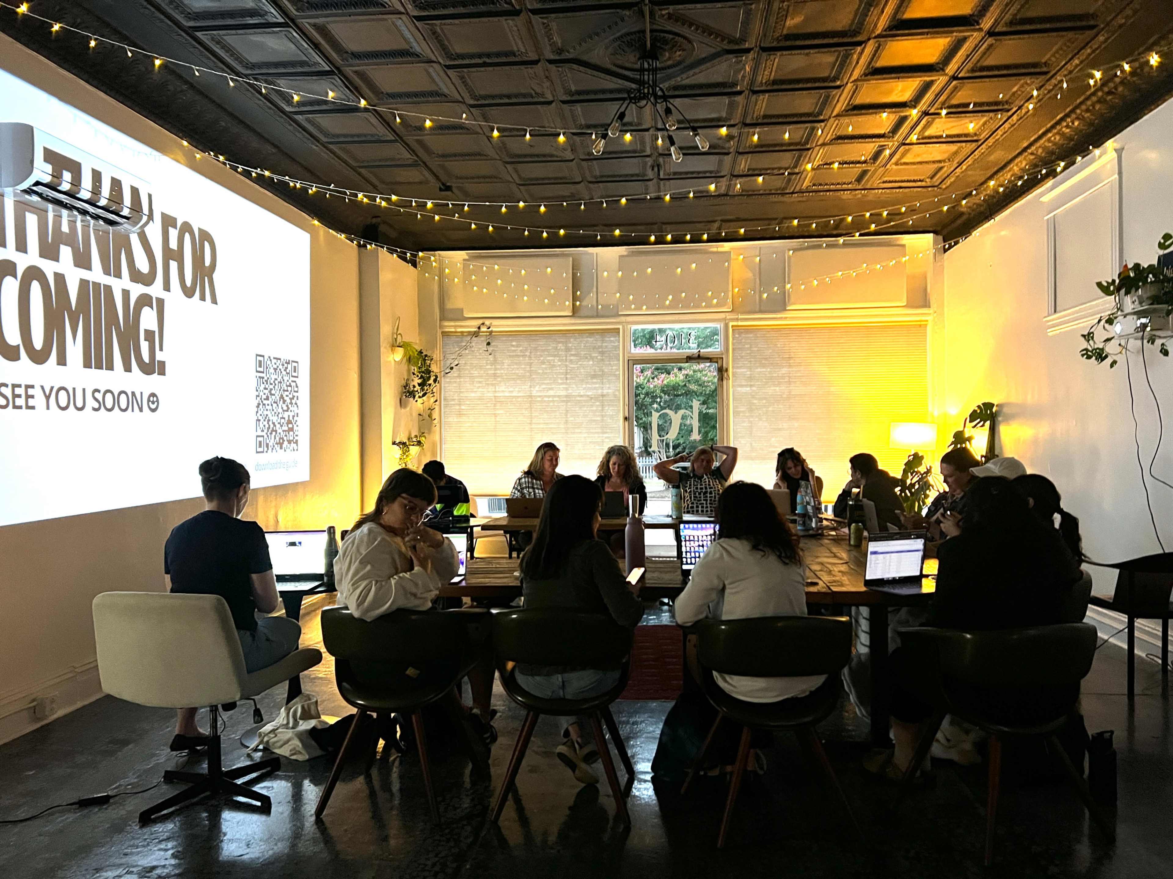 A group of people is seated around a long table in a cozy, well-lit meeting room, with a large screen displaying a "Thanks for Coming!" message.