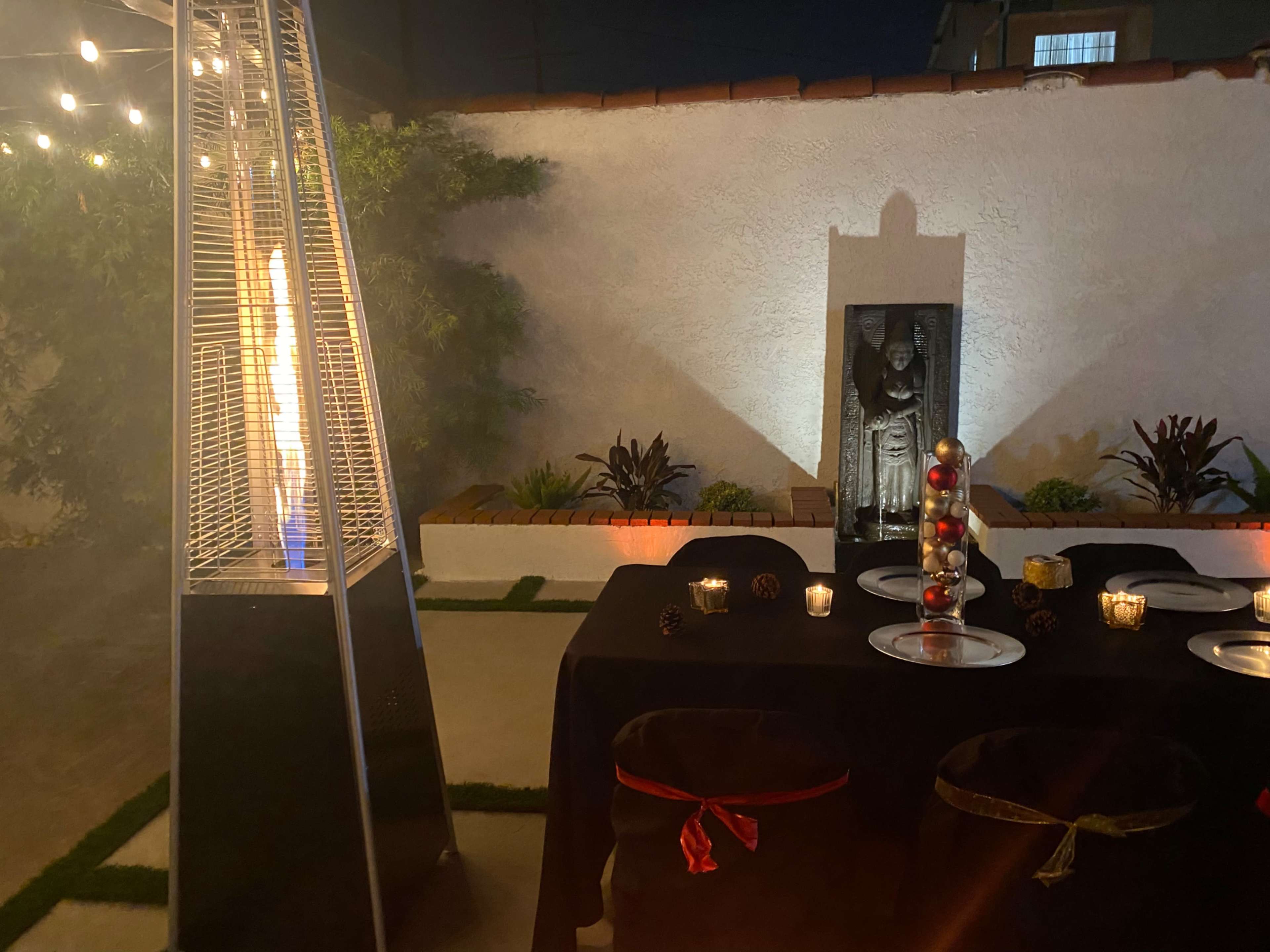 An outdoor dining area features a table set with black chairs, illuminated candles, and a patio heater beside a decorative statue against a white wall.