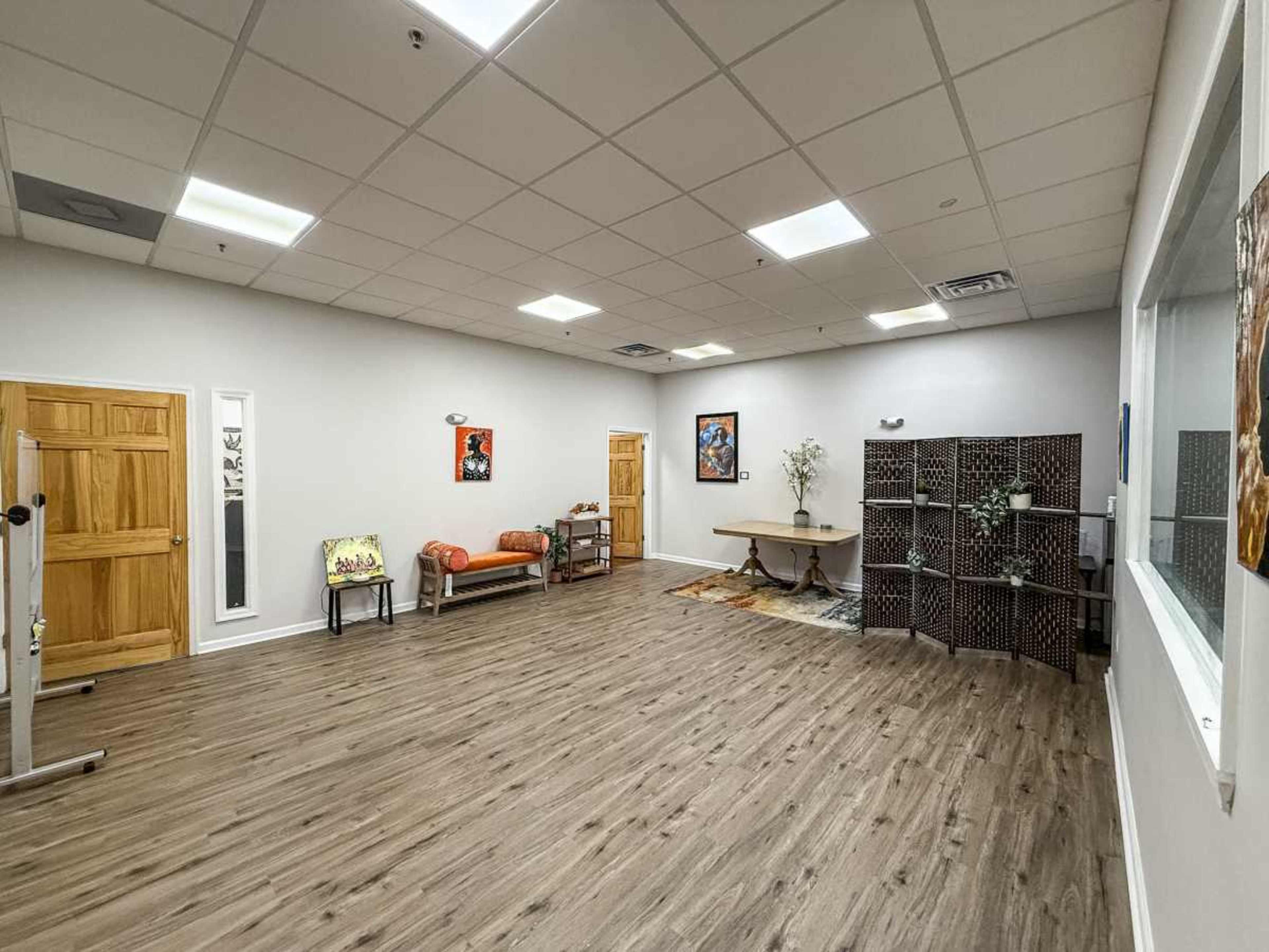 The image shows a spacious, well-lit room with wooden flooring, featuring a round table, a sofa, a decorative partition, and a few pieces of artwork on the walls.
