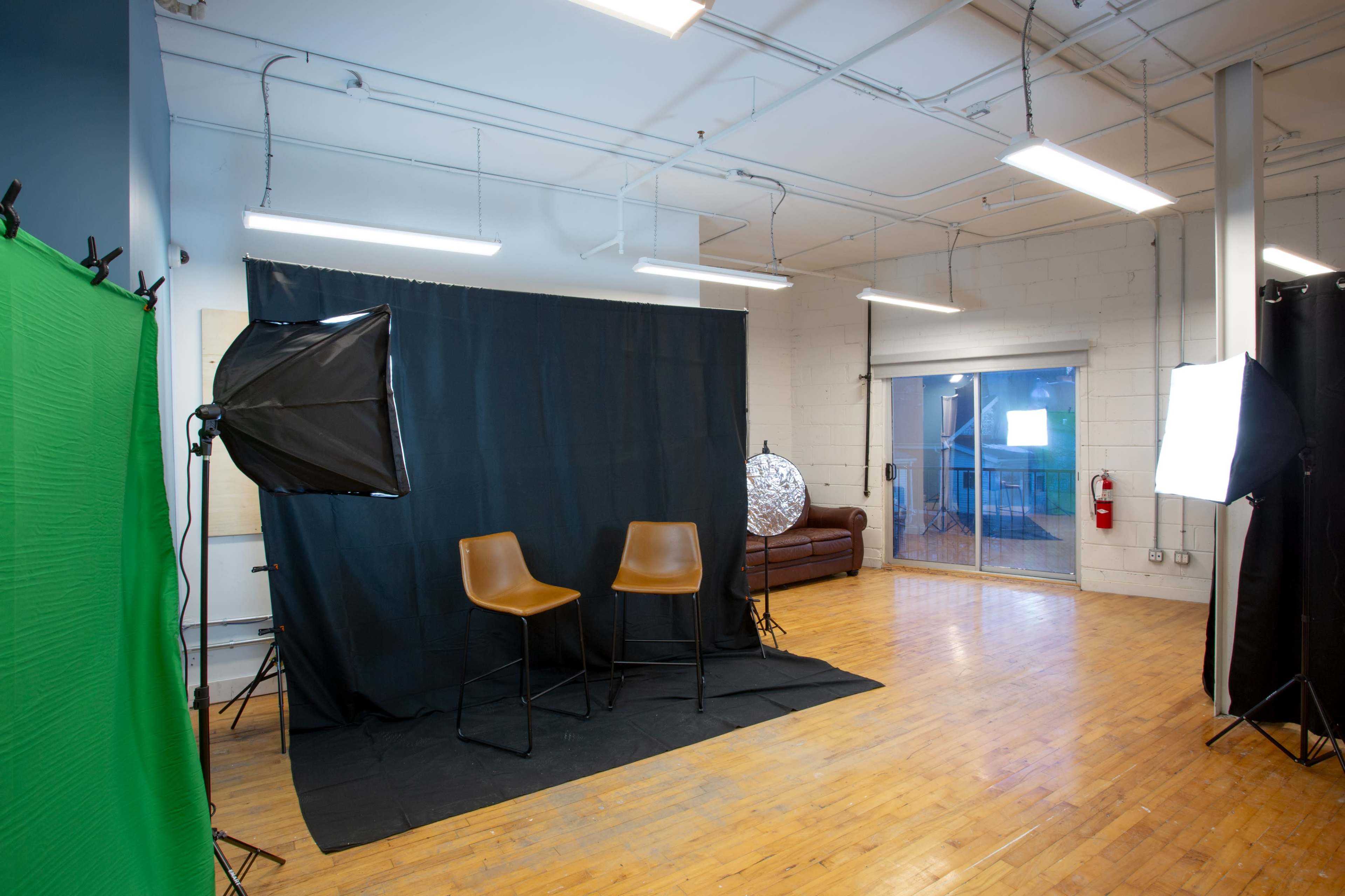 A studio setup with two brown chairs on a black mat, green and black backdrops, and lighting equipment arranged around the space.