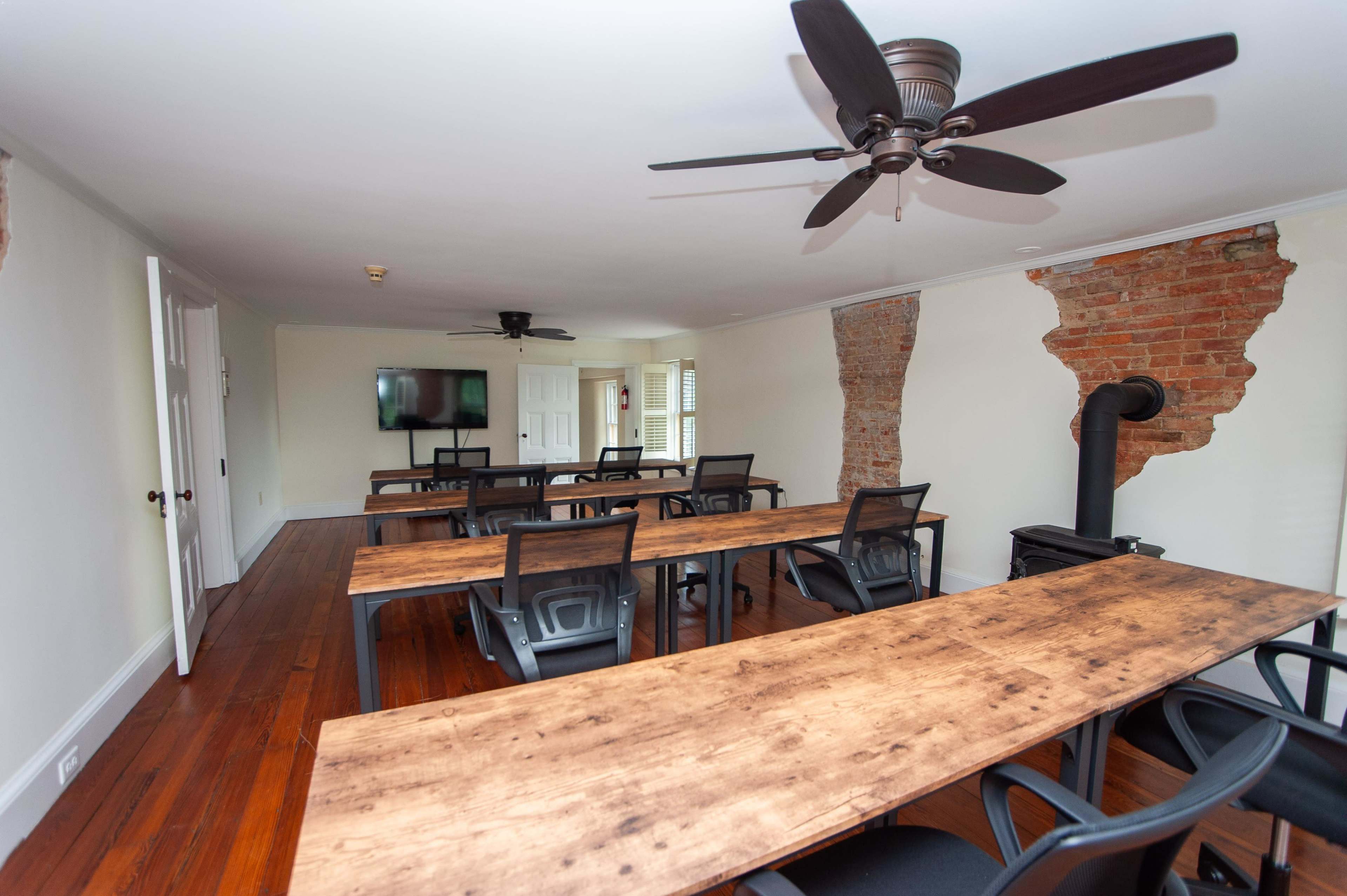 A conference room with wooden tables, black chairs, a television on the wall, and exposed brick accents.