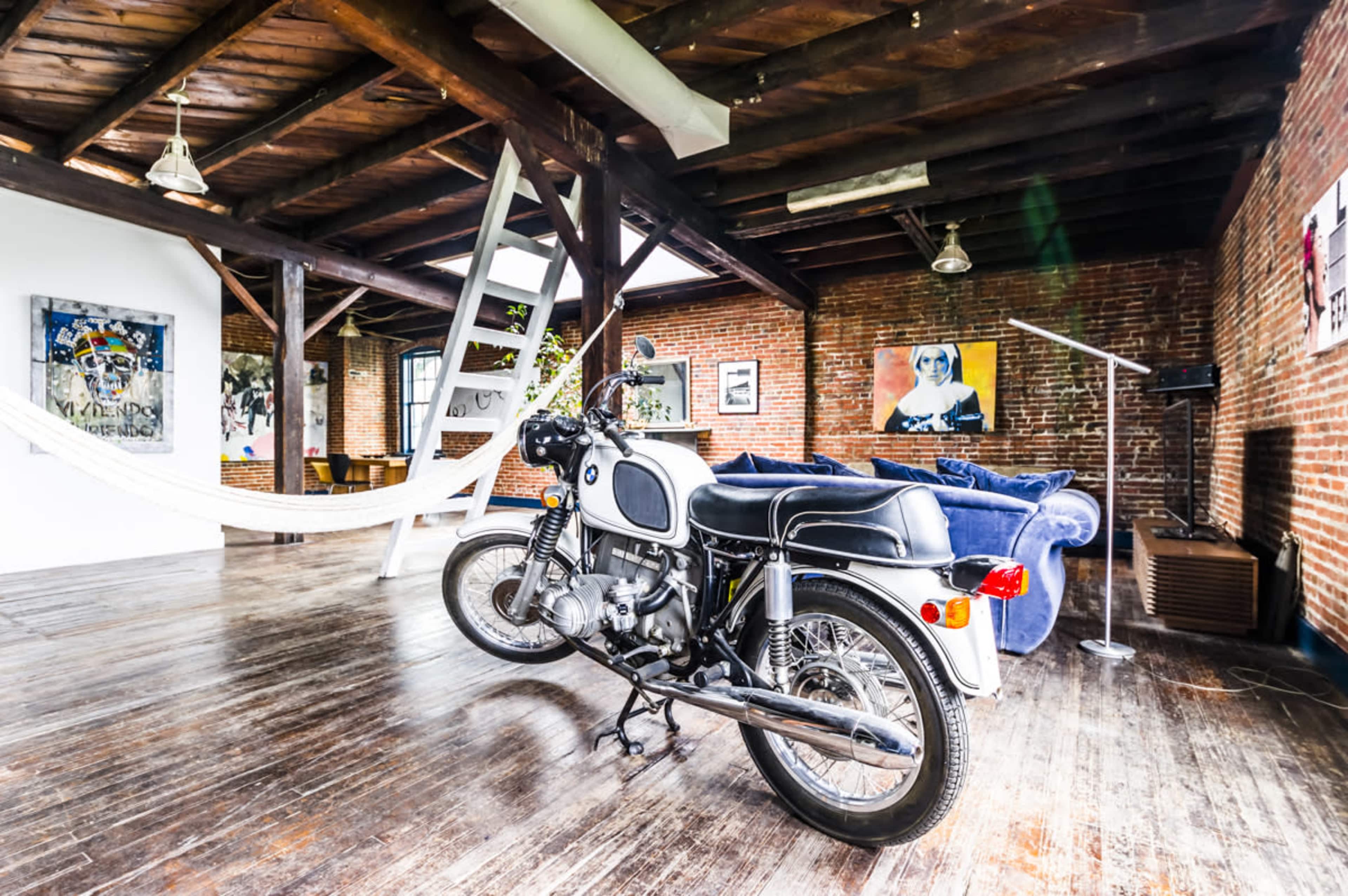 A vintage motorcycle is parked on a wooden floor in an open-concept loft with exposed brick walls and modern furnishings.
