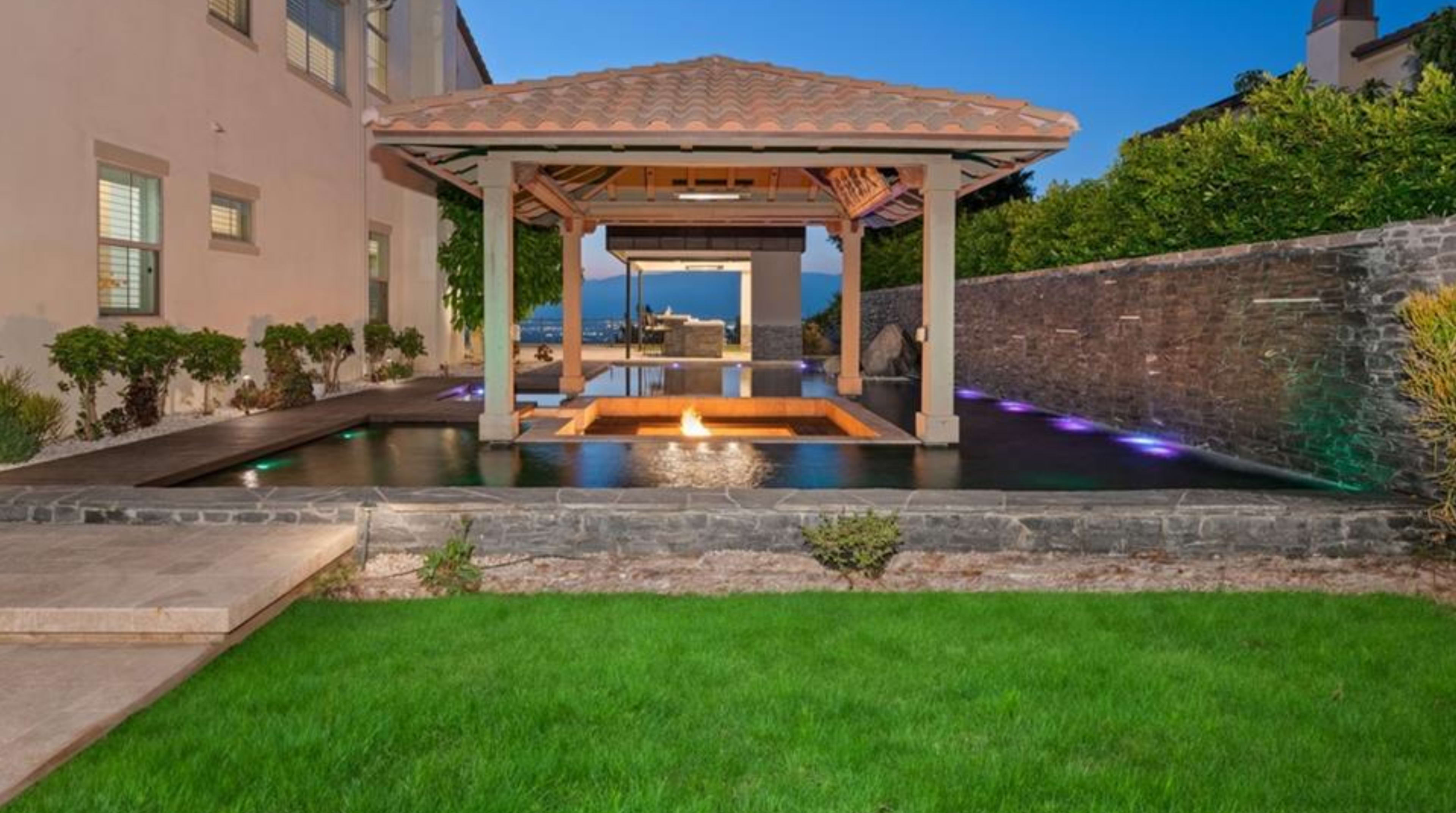 The image shows a landscaped outdoor area featuring a gazebo over a rectangular pool with a wooden deck and fire feature, surrounded by stone walls and manicured grass.