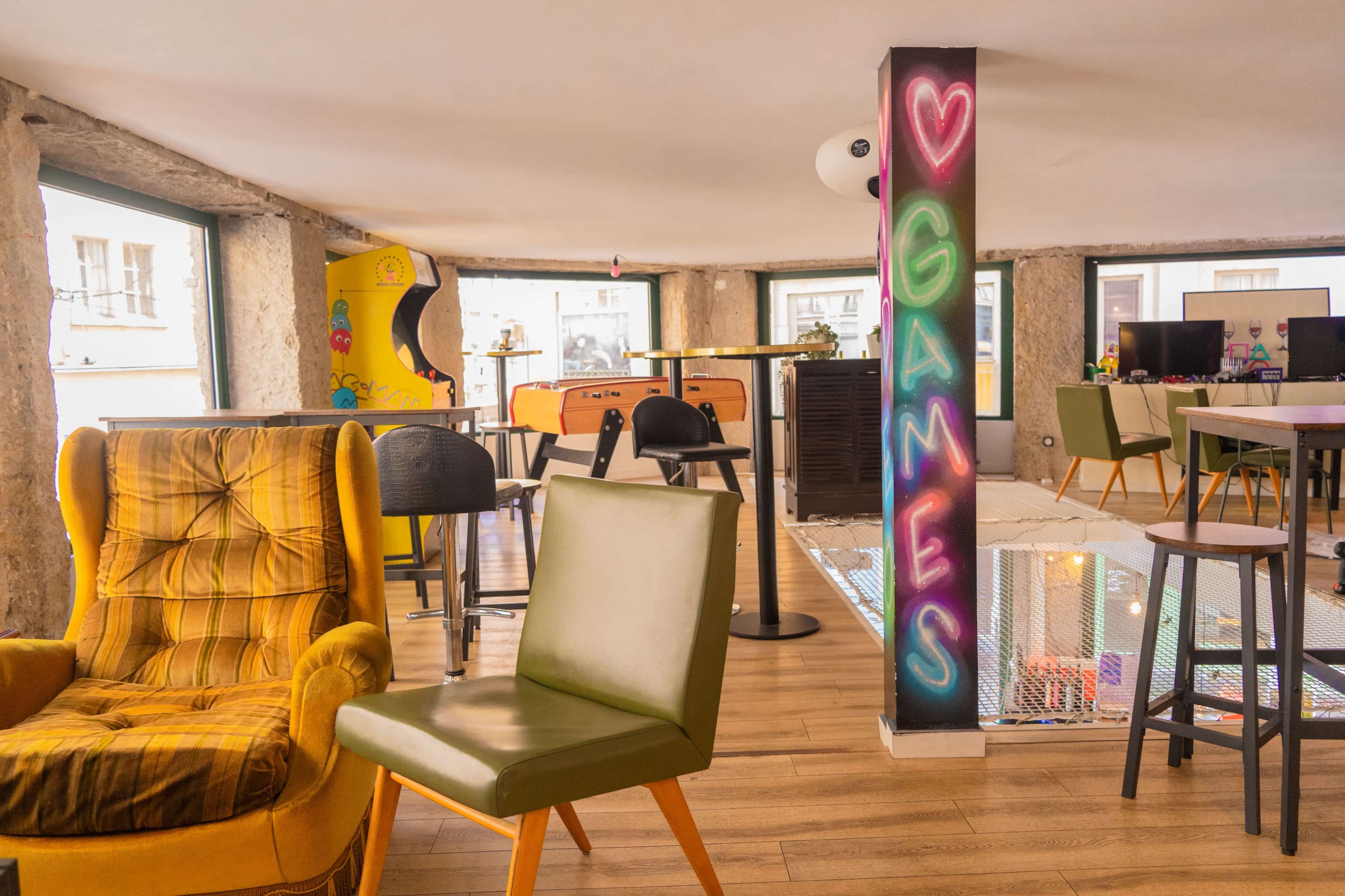 The image shows an indoor lounge area featuring colorful furniture, arcade machines, and a vibrant "GAMES" mural on a column.