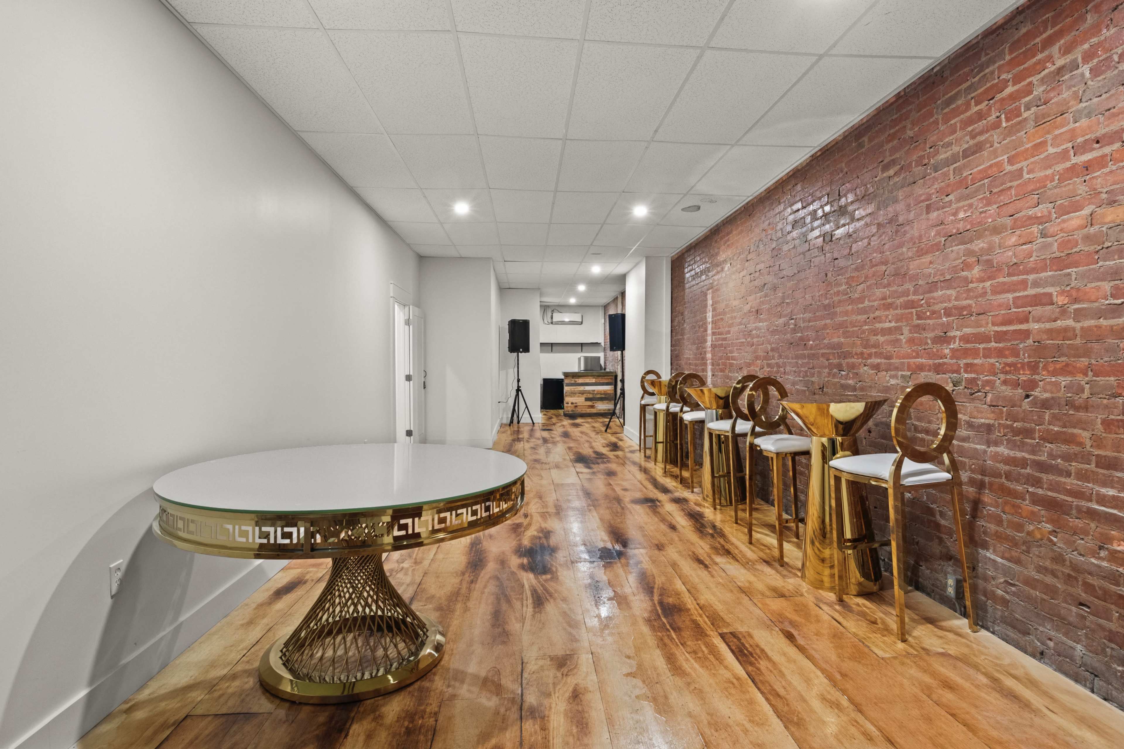 A modern interior space with a round table and gold-accented chairs along a brick wall, featuring a polished wooden floor and ceiling lights.